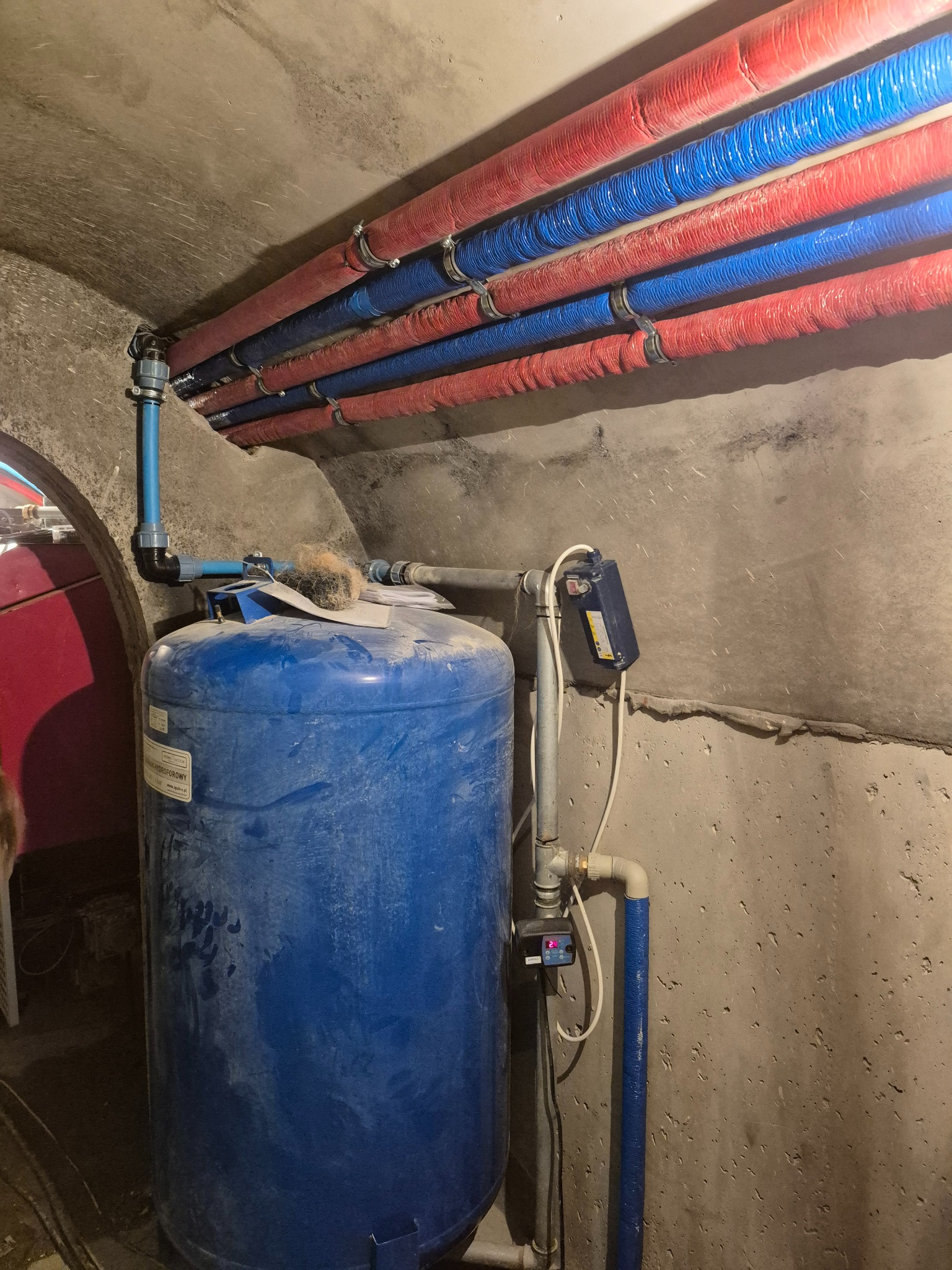 Hydrofor z osprzętem i rurami w izolacji, zamontowany w pomieszczeniu o surowych, betonowych ścianach. Rury w kolorach niebieskim i czerwonym biegną wzdłuż sufitu.