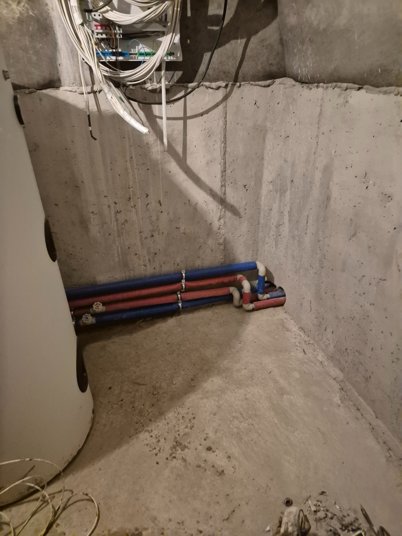 Instalacja rur wodnych (czerwone i niebieskie) w betonowej piwnicy, obok białego zbiornika. Widoczne kable elektryczne pod sufitem. Surowy stan wykończenia.