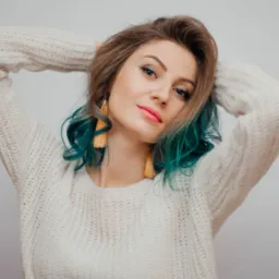 Portret młodej kobiety z turkusowymi końcówkami włosów, w kremowym swetrze, pozującej z rękami uniesionymi za głową, na jasnym tle.