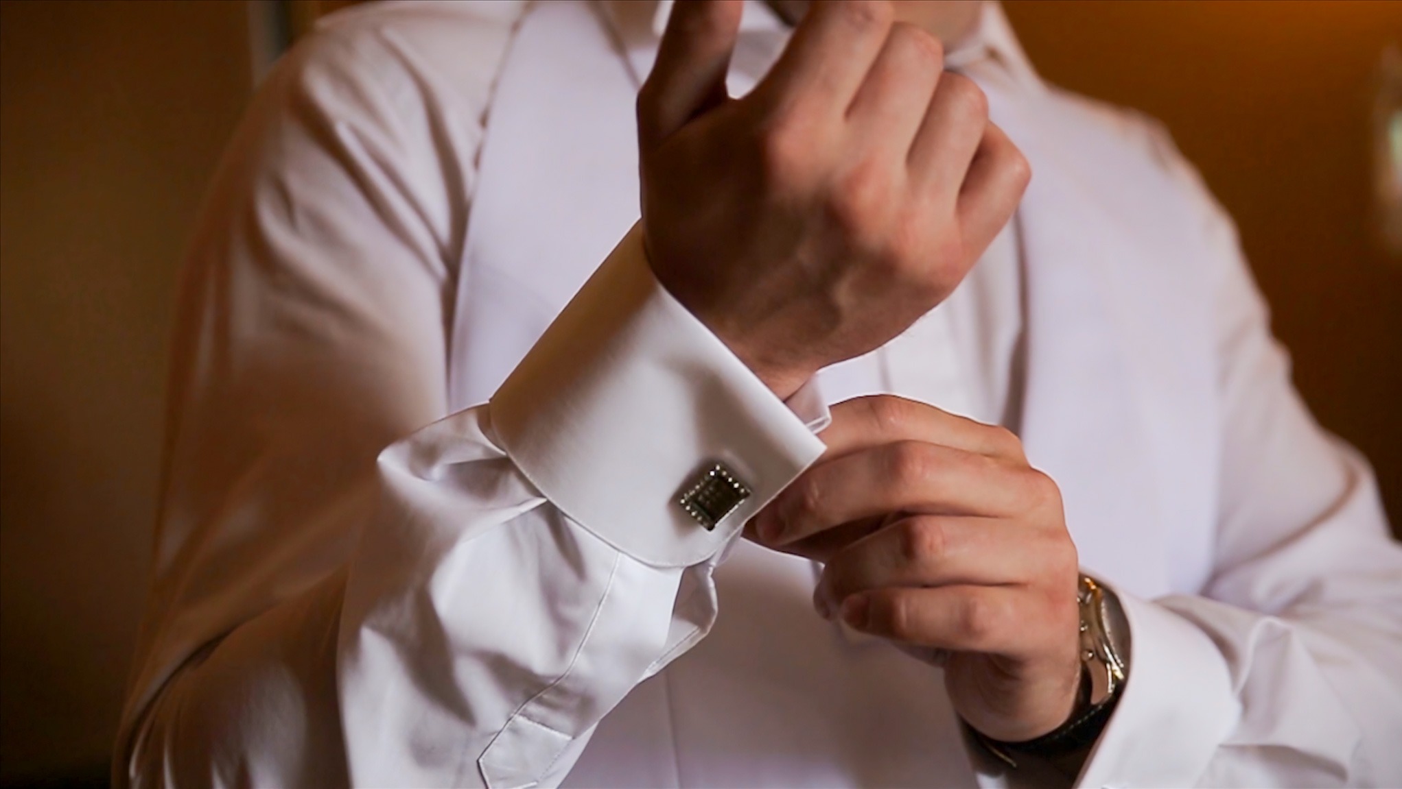 Mężczyzna w białej koszuli i kamizelce zapina ozdobny mankiet z kwadratowym spinką, widoczny zegarek na nadgarstku.