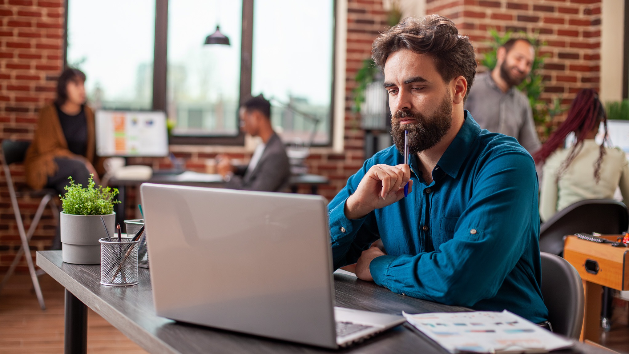 Mężczyzna z brodą w niebieskiej koszuli zamyślony przed laptopem w biurze typu open space, trzymając długopis przy ustach, w tle inni pracownicy i ceglana ściana.