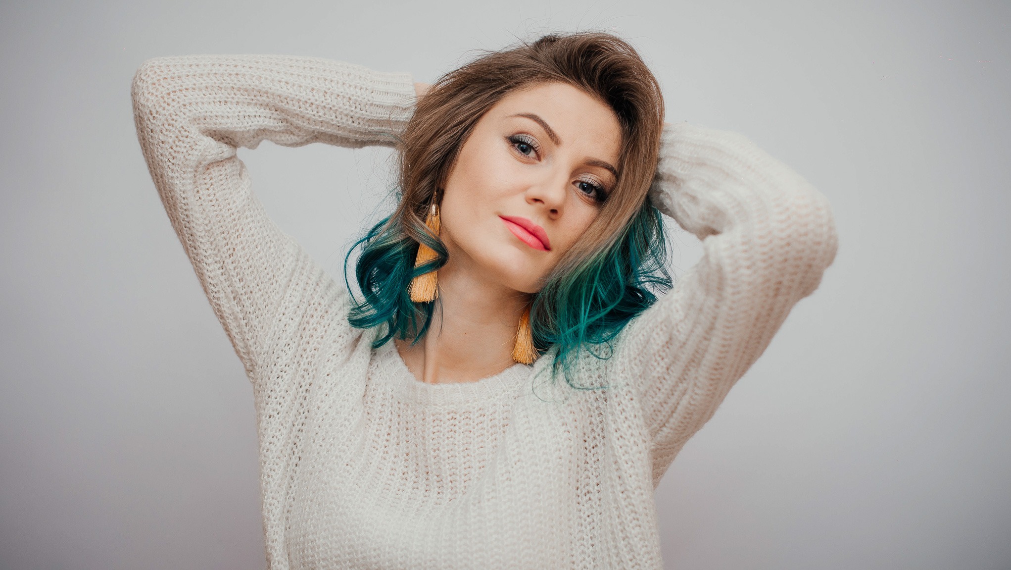 Portret młodej kobiety z turkusowymi końcówkami włosów, w kremowym swetrze, pozującej z rękami uniesionymi za głową, na jasnym tle.