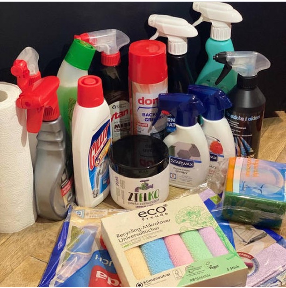 Zestaw różnych środków czyszczących i akcesoriów do sprzątania, w tym spraye, płyny, pasty, gąbki i ściereczki z mikrofibry, ułożone na drewnianej powierzchni.