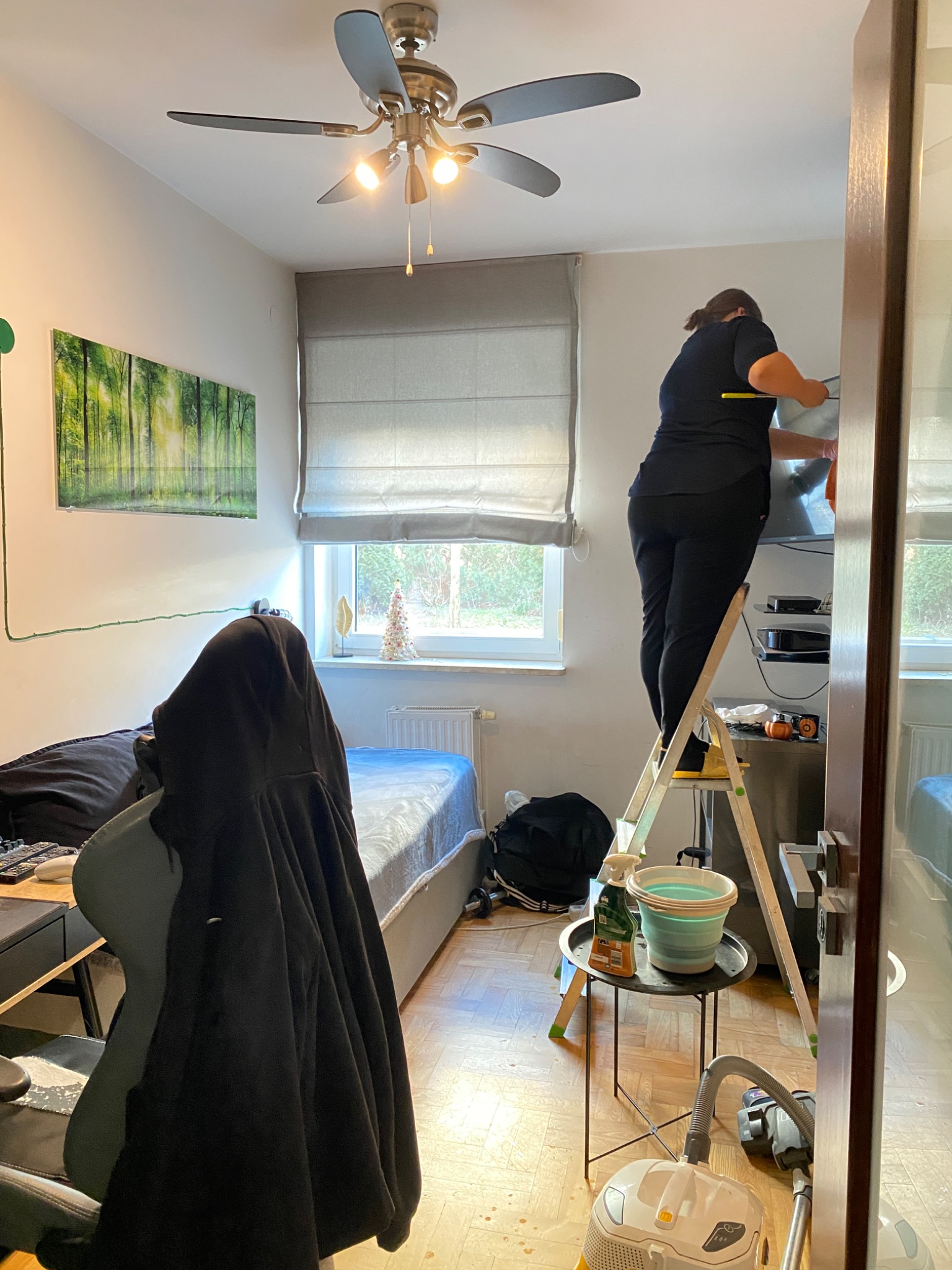 Osoba na drabine czyści telewizor w pokoju, widoczne wiadro, środek czyszczący, odkurzacz i elementy wyposażenia pokoju.