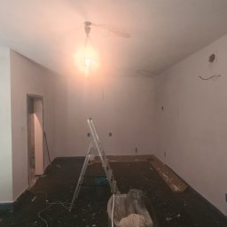 K-BUD Łukasz Kruczkowski - Wnętrze po gruntowaniu ścian na biało, widoczna drabina, zabezpieczona podłoga i surowa żarówka. Przygotowanie do malowania.