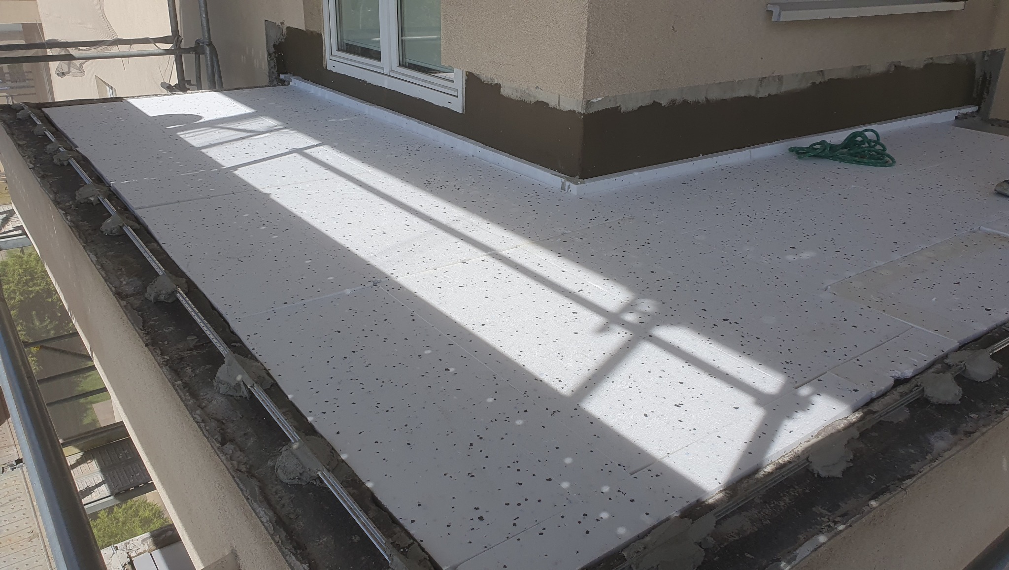 Białe płyty izolacyjne na balkonie w trakcie montażu, widoczne listwy przyścienne i fragment elewacji z oknem, cień rzucany przez barierki.