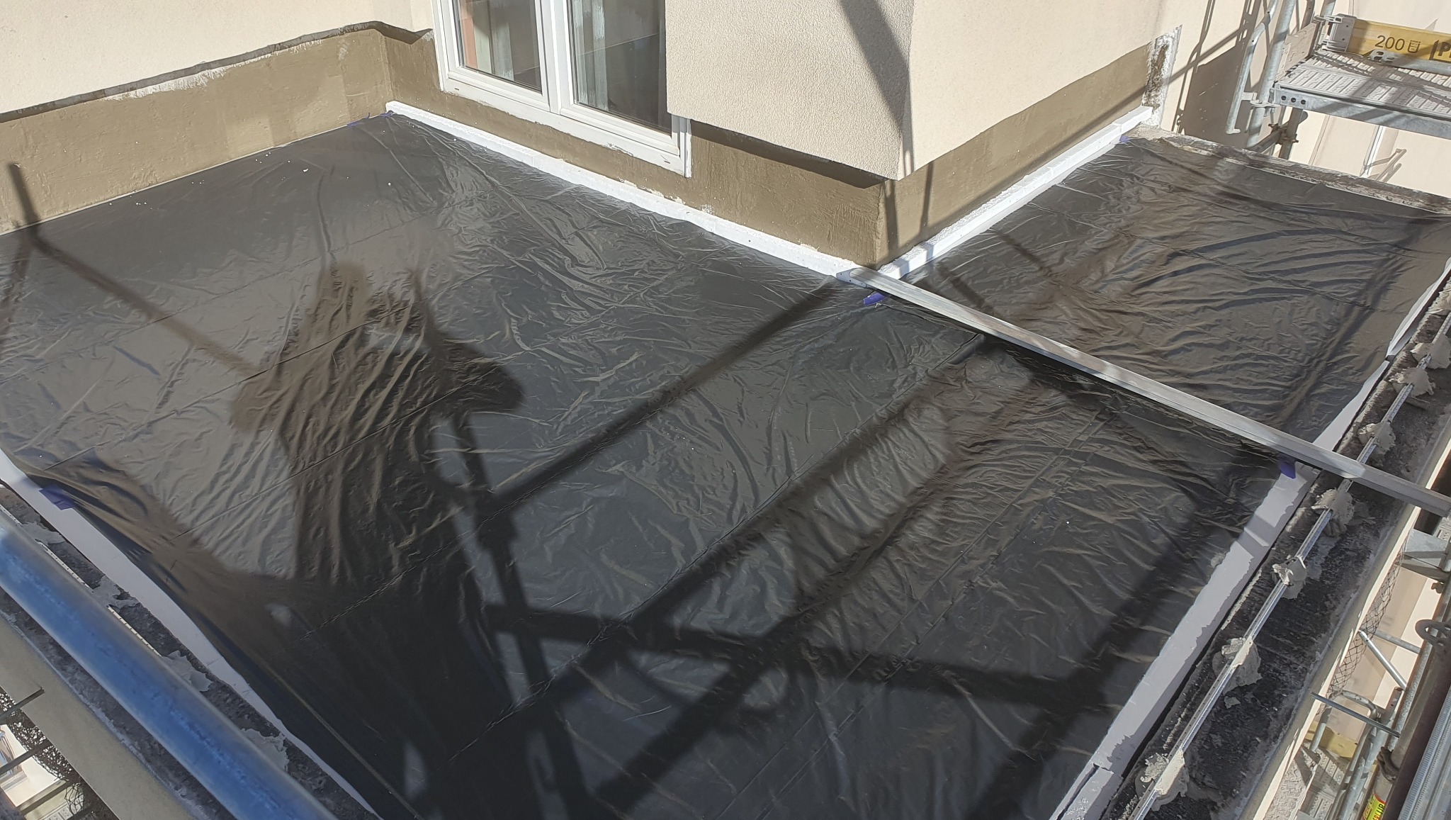 Przygotowanie balkonu do prac wykończeniowych: zabezpieczona folią powierzchnia, świeżo pomalowane obrzeża i poziomica na rusztowaniu.