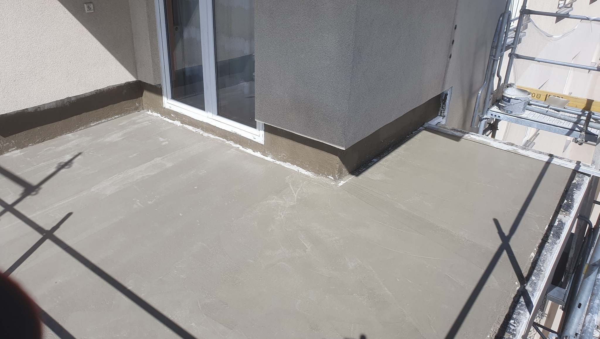 Wyrównana, szara wylewka na balkonie z widoczną stolarką okienną i fragmentem elewacji. Na rusztowaniu stoi wiadro z farbą.