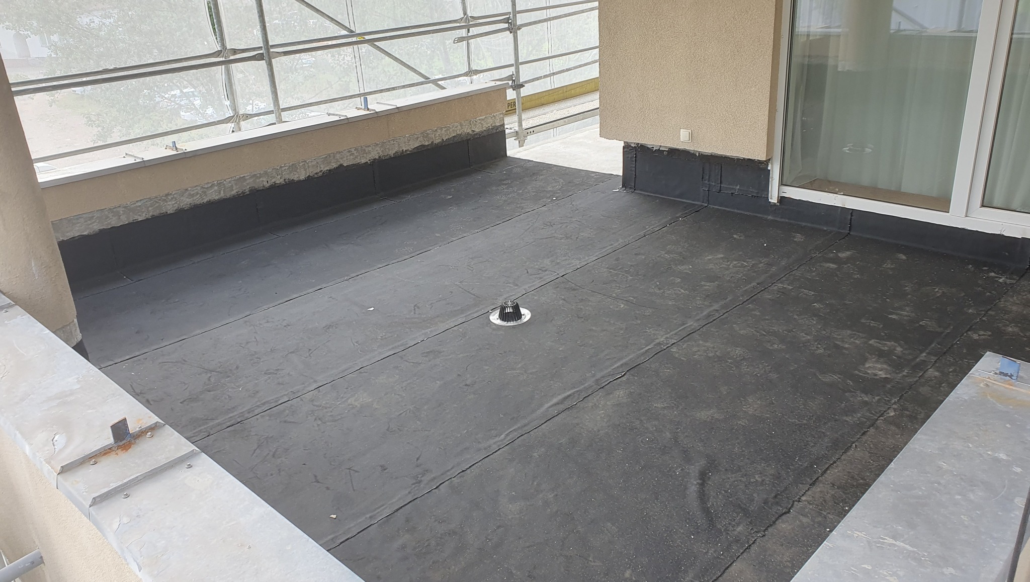 Balkon po hydroizolacji papą termozgrzewalną, widoczny odpływ liniowy, rusztowania w tle. Ściany otynkowane, okno balkonowe. Prace wykończeniowe na balkonie.