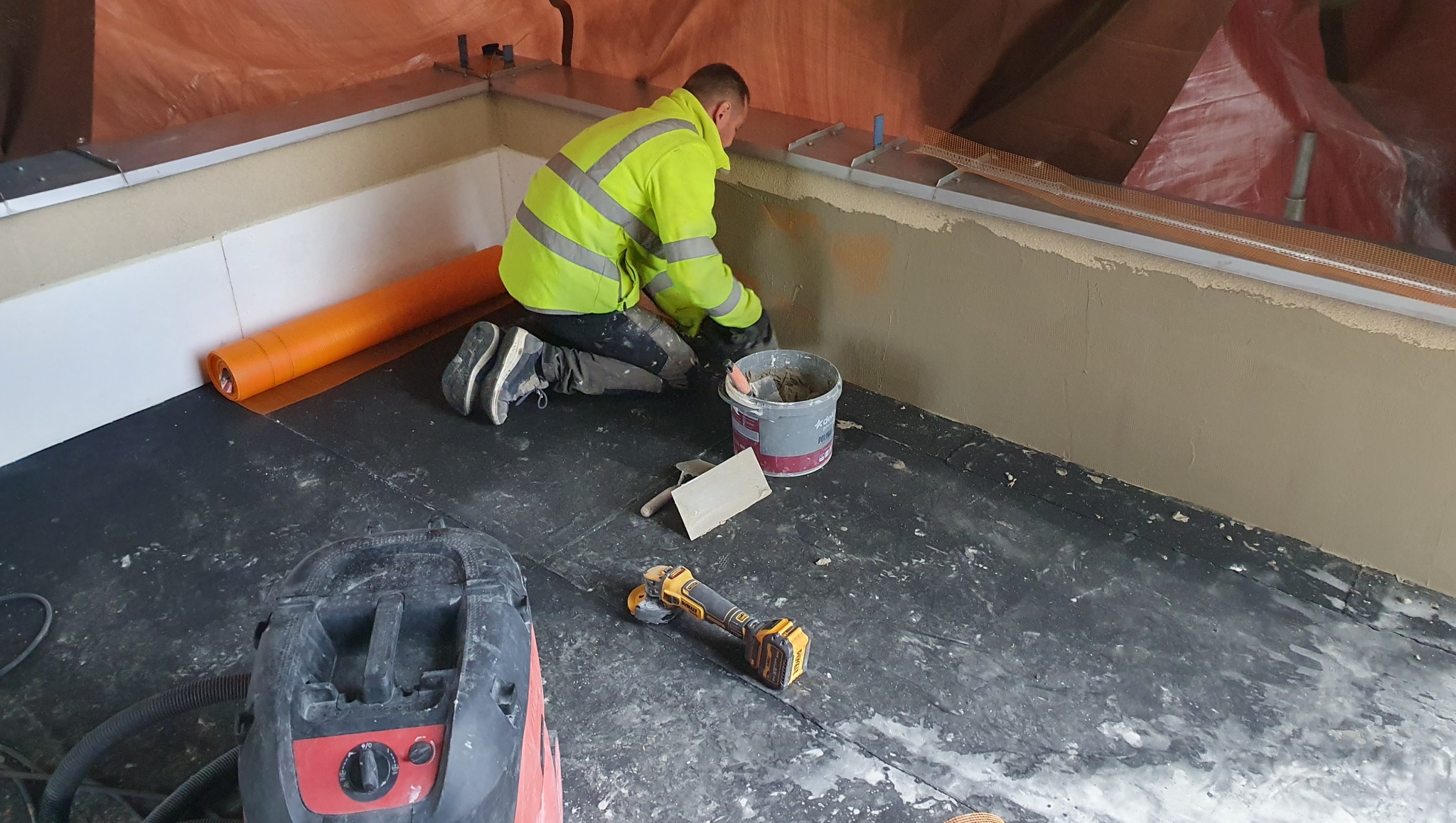 Pracownik w żółtej kurtce szpachluje ścianę w pomieszczeniu. Widoczne narzędzia, wiadro z zaprawą, odkurzacz i pomarańczowa rolka.