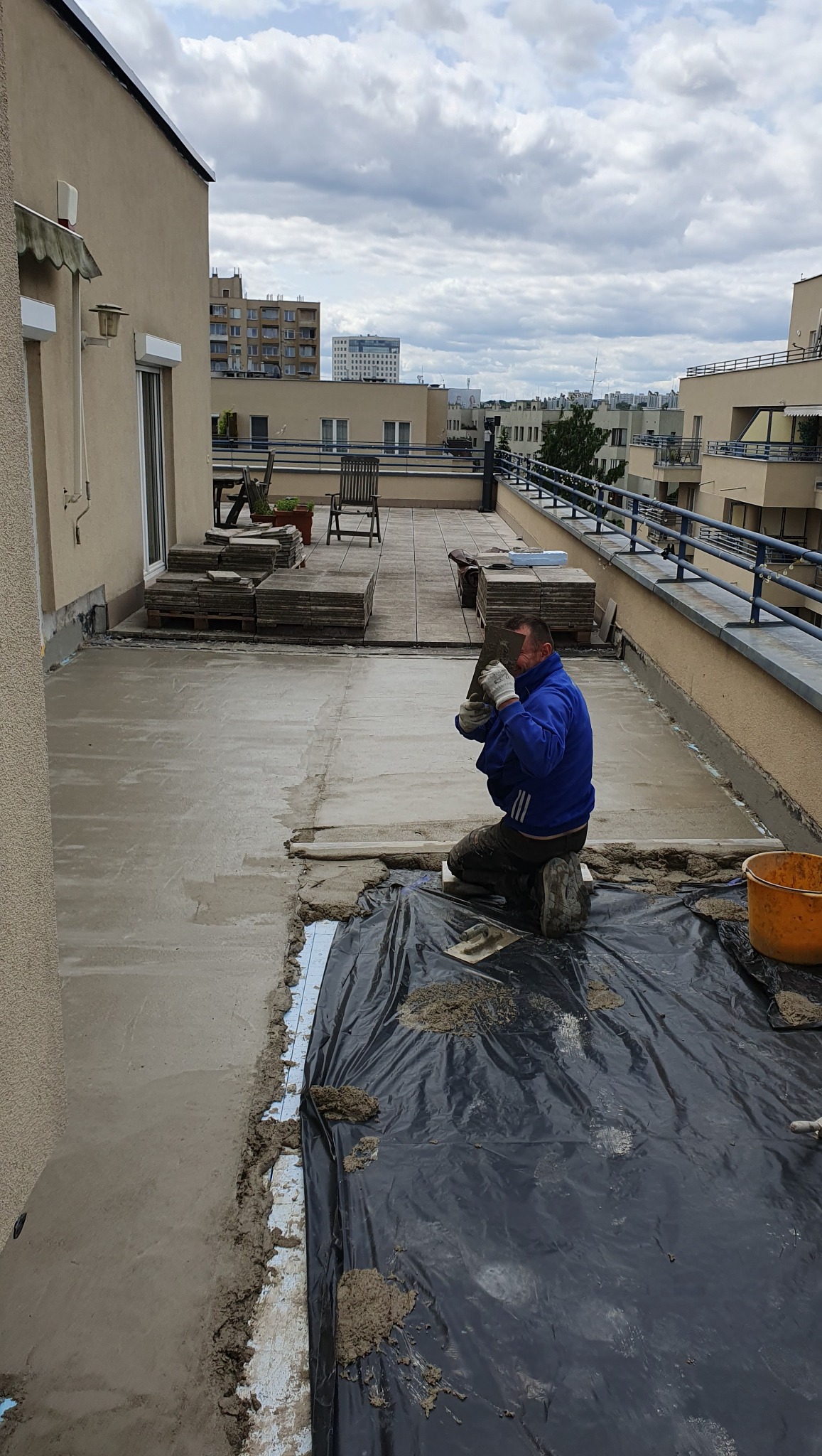 Pracownik w niebieskiej bluzie szpachluje taras na dachu budynku. Część powierzchni wygładzona, reszta przykryta folią. W tle budynki miejskie i pochmurne niebo.