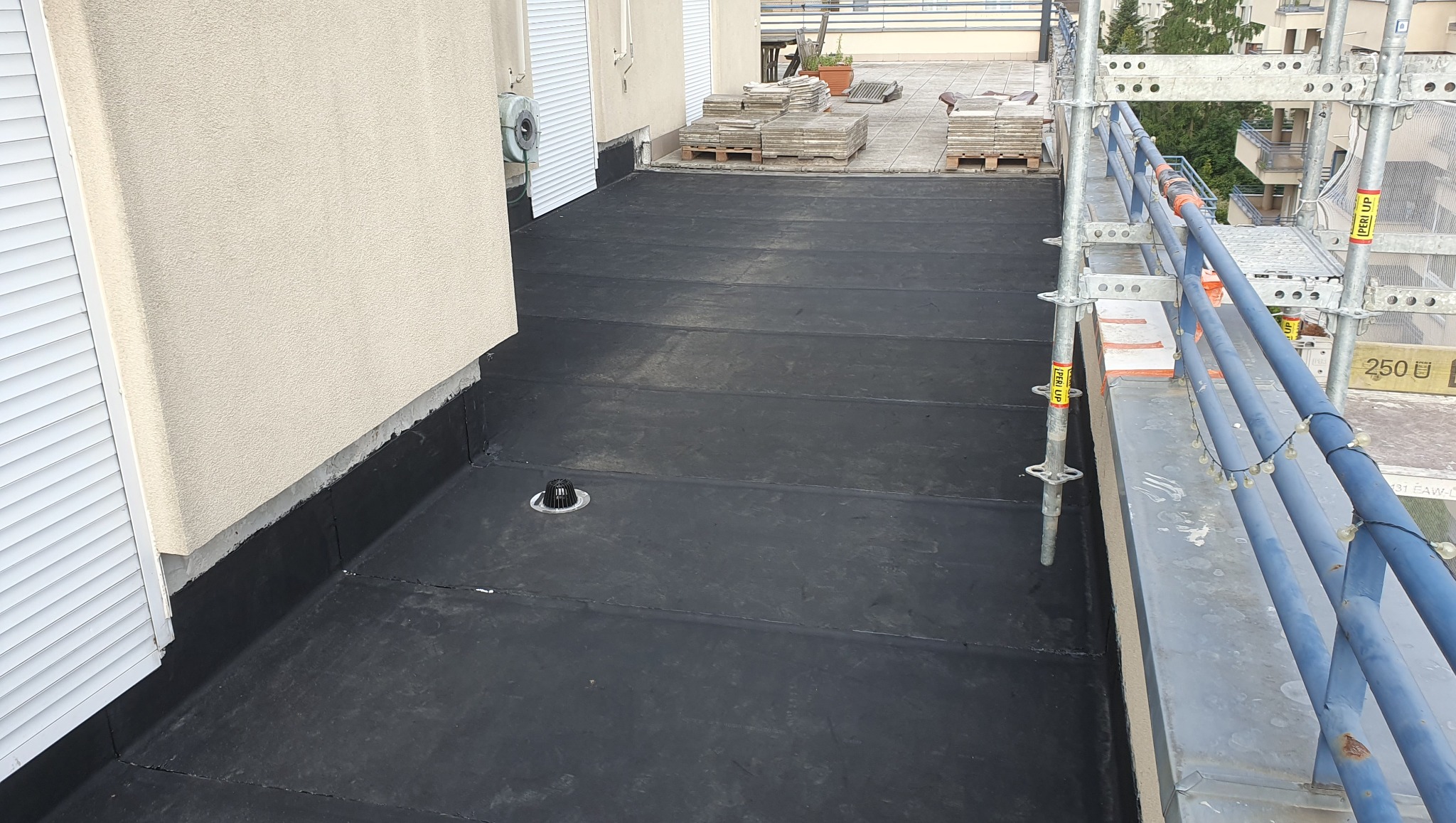 Wykończony dach pokryty czarną papą termozgrzewalną, widoczny system odprowadzania wody, rusztowanie i fragment elewacji budynku z żaluzjami.