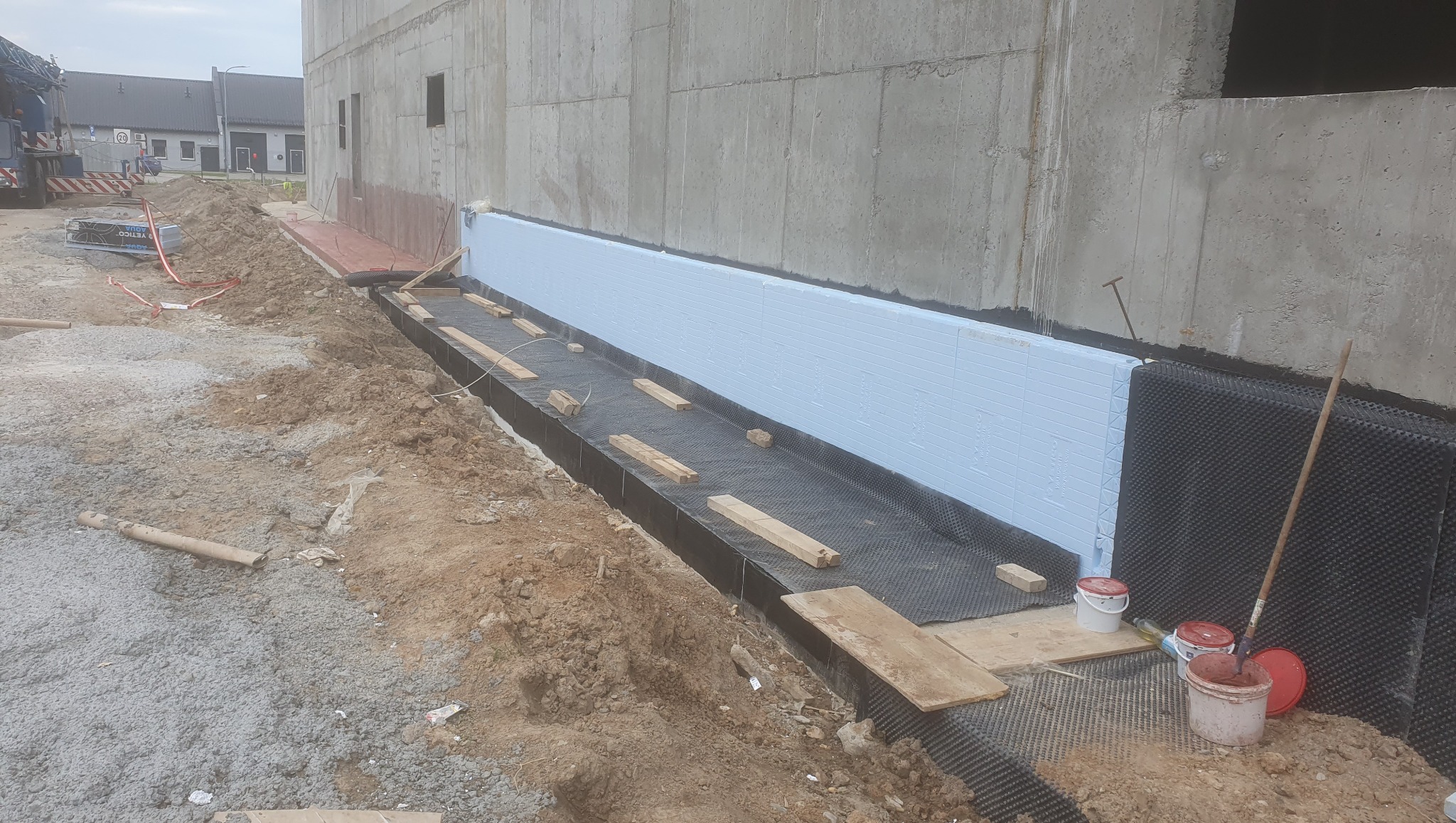 Izolacja fundamentów budynku z użyciem styropianu i folii kubełkowej, widoczne narzędzia i materiały budowlane, teren budowy.