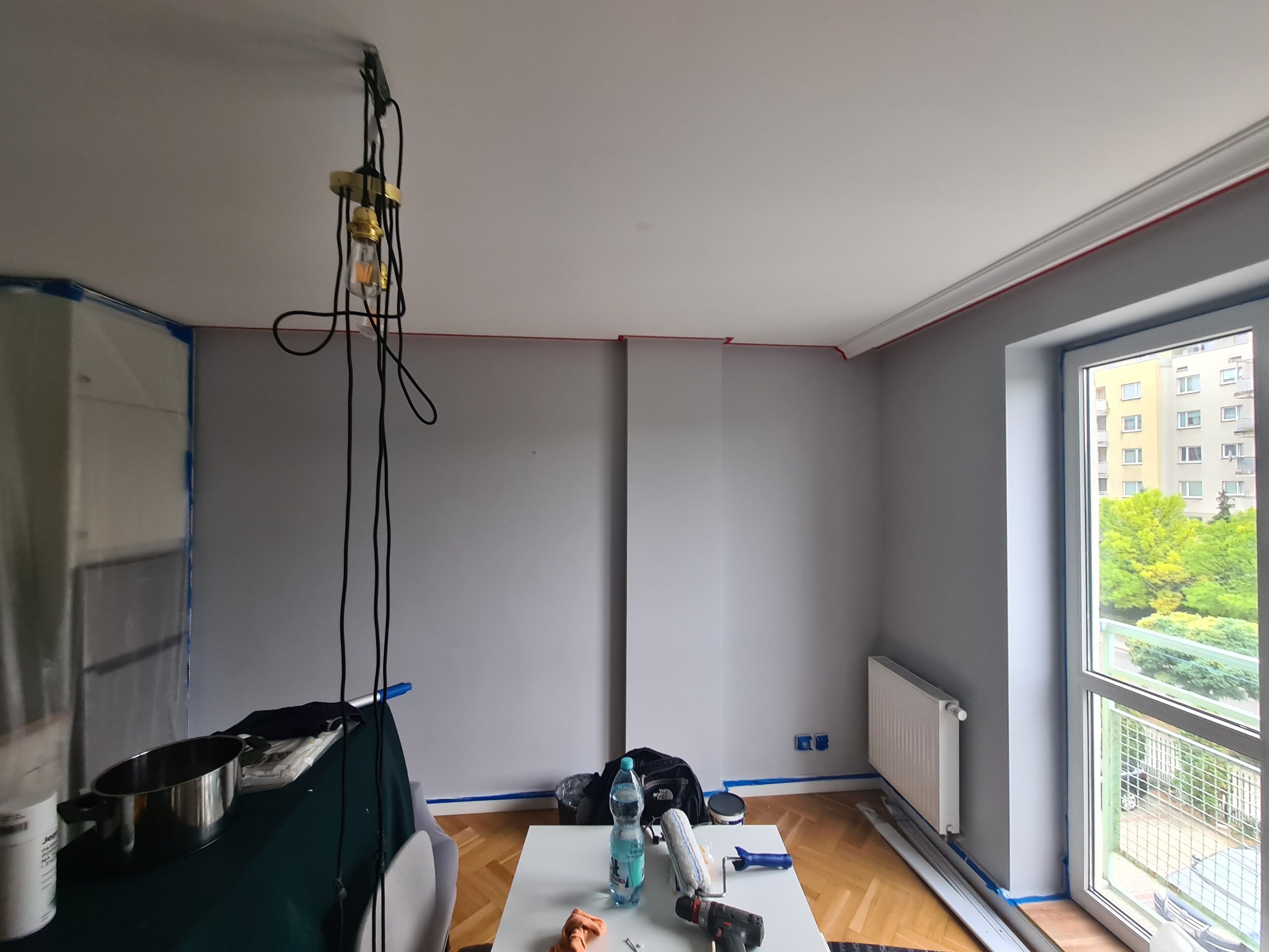 Pokój po malowaniu na szaro, z zabezpieczonymi listwami i oknami taśmą malarską. Widoczne narzędzia malarskie na stole i lampa wisząca. Parkiet na podłodze.