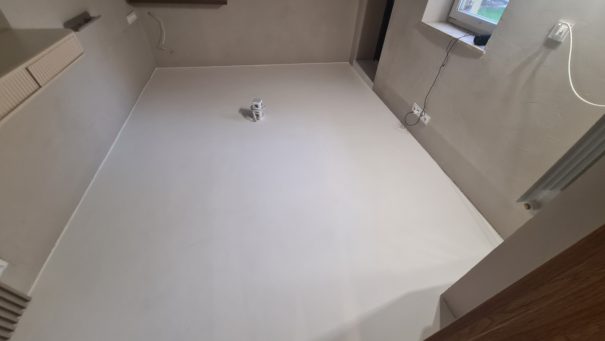 Jasne wnętrze po wylaniu żywicy na podłogę. Widoczne gładkie, jednolite wykończenie podłogi, ściany w odcieniach beżu, okno i wystające kable elektryczne.