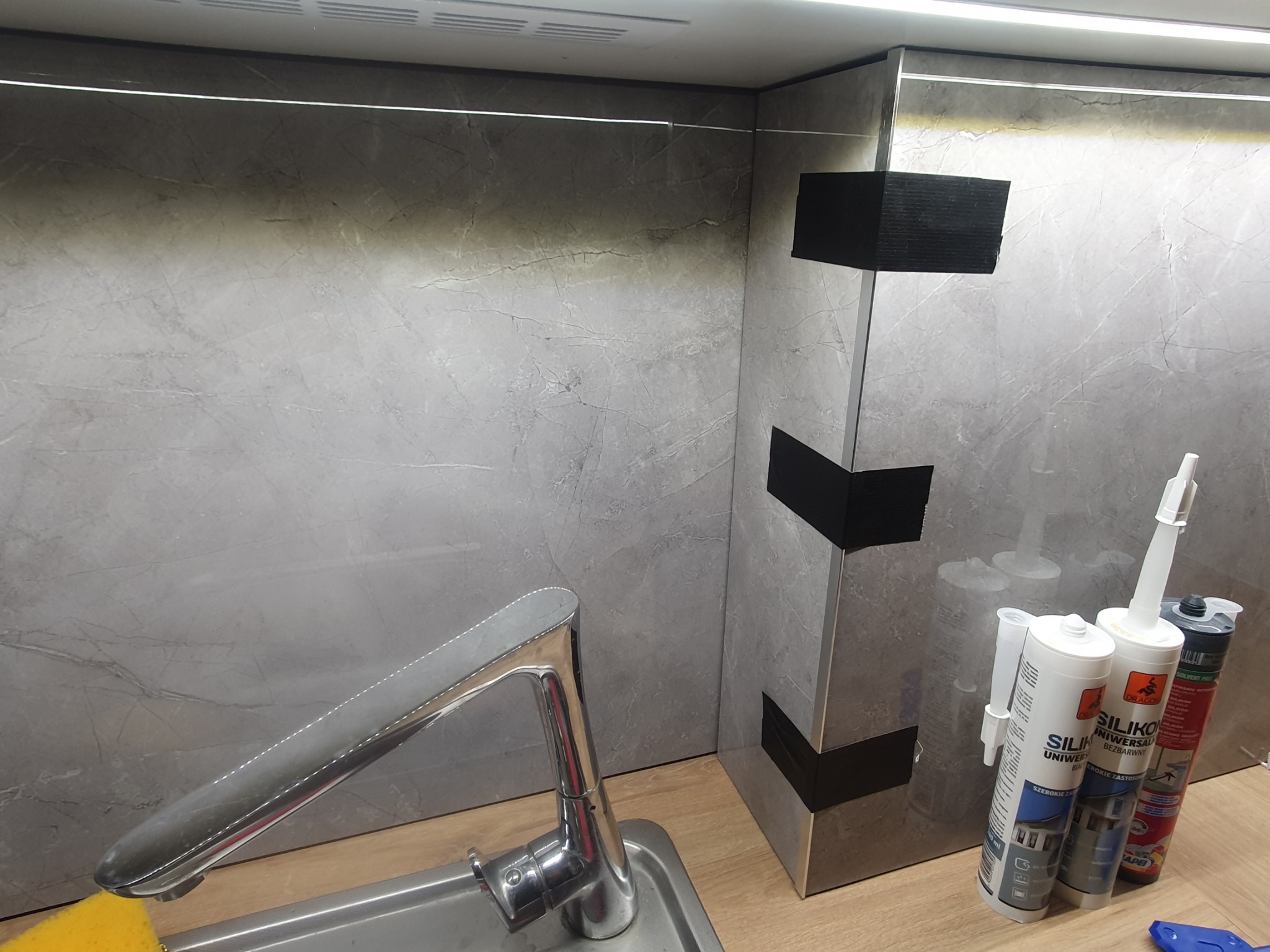 Wykończona ściana w kuchni z szarych płytek imitujących marmur, widoczny kran, zlew i silikony budowlane. Fragment blatu z drewna.