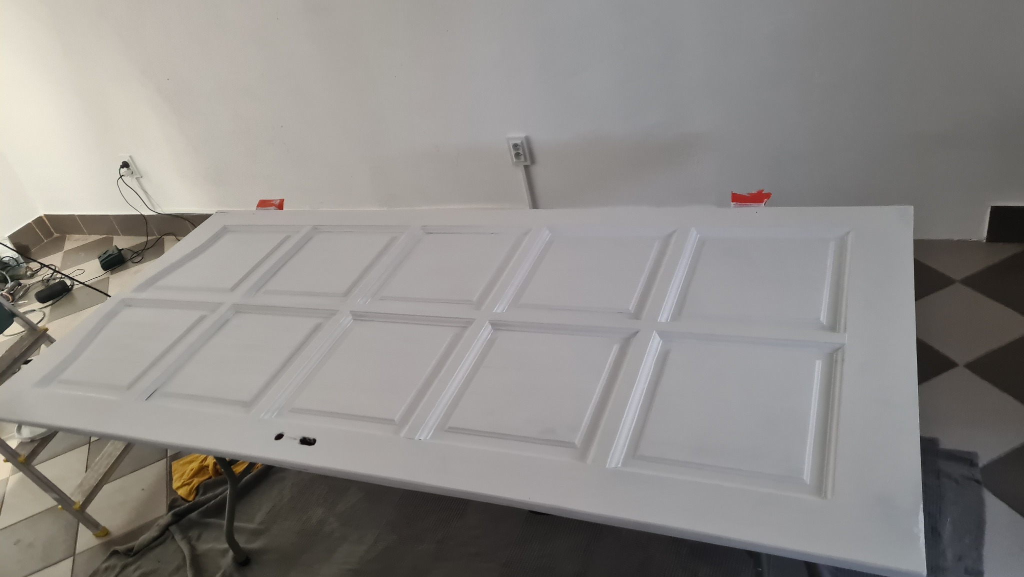 Białe, świeżo pomalowane drzwi z panelami, leżące na kozłach malarskich w jasnym pomieszczeniu. Widoczne narzędzia malarskie i gniazdko elektryczne na ścianie.