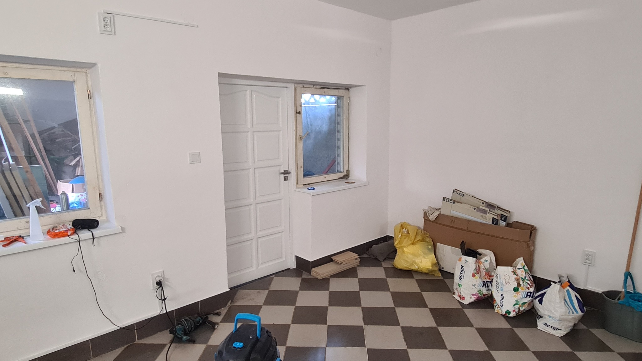 Wnętrze po malowaniu na biało: ściany, drzwi i okno odświeżone, na podłodze szachownica, widoczne narzędzia i worki po materiałach budowlanych.
