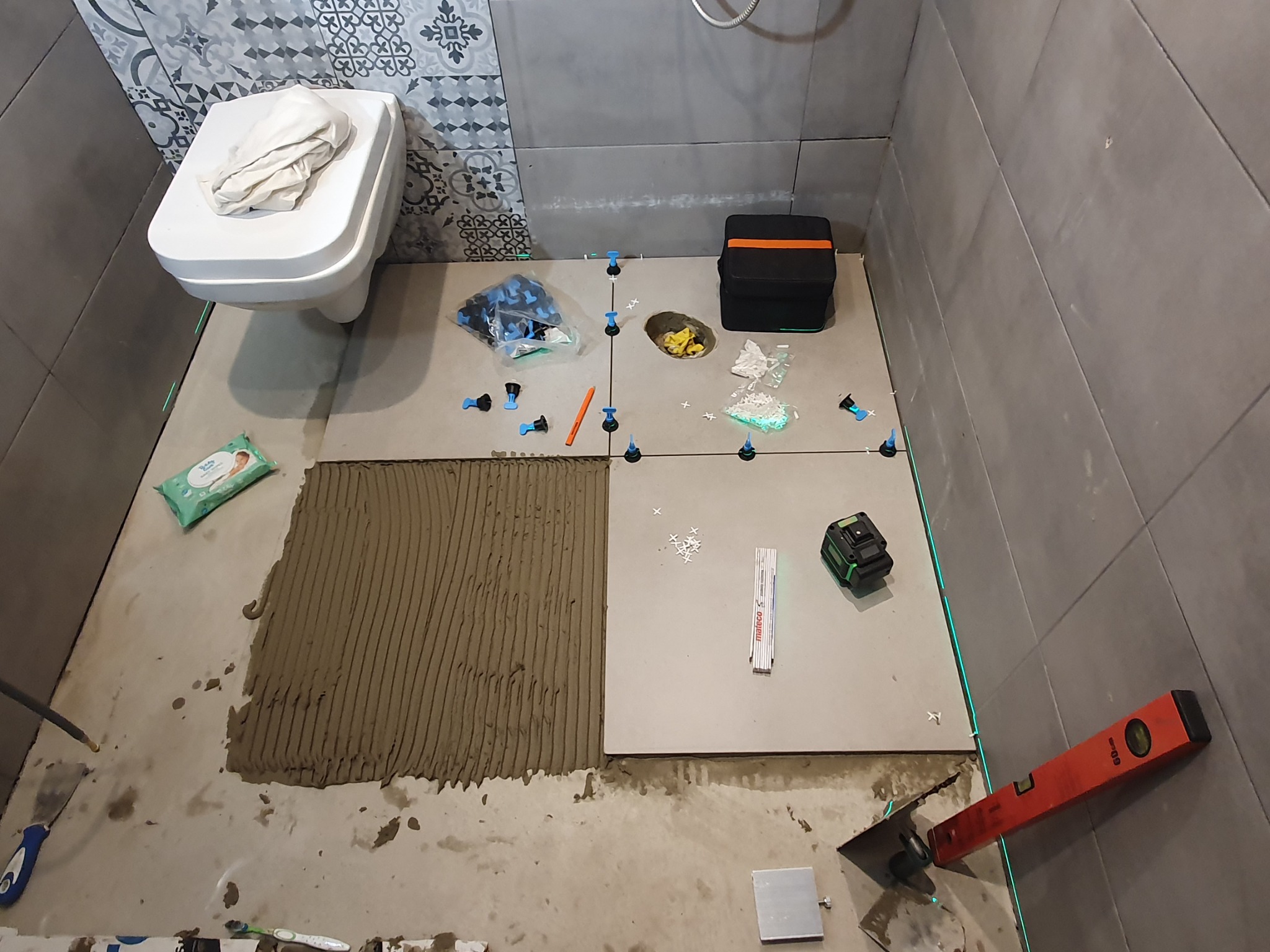 Układanie płytek podłogowych w łazience z użyciem poziomicy laserowej i systemu poziomowania. Widoczna nałożona zaprawa i płytki w trakcie montażu.