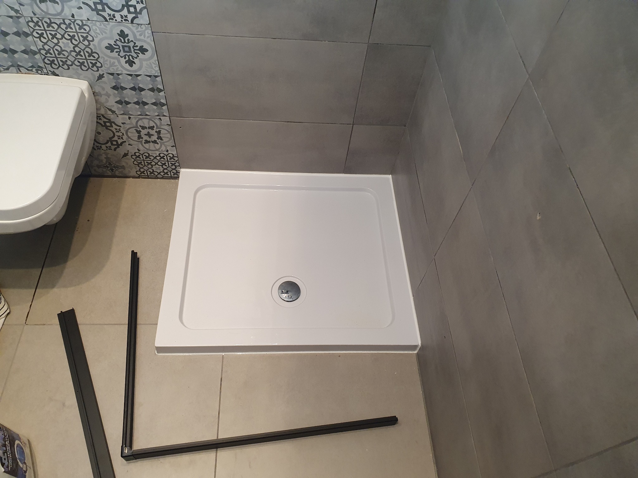 Biały brodzik z odpływem w rogu łazienki z szarymi płytkami i dekoracyjnym pasem. Elementy czarnej ramy kabiny prysznicowej leżą na podłodze.