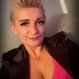 Portret kobiety z krótkimi blond włosami, w czarnej marynarce i różowej bluzce, z delikatnym uśmiechem i subtelną biżuterią.