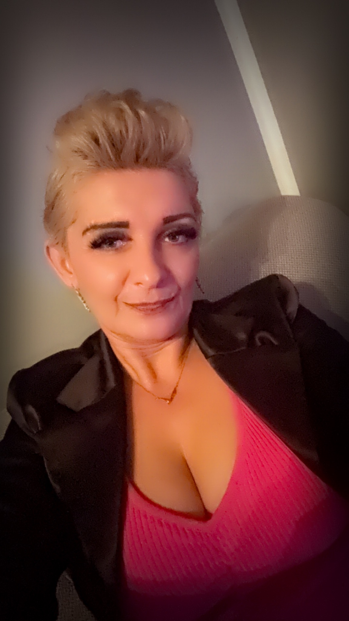 Portret kobiety z krótkimi blond włosami, w czarnej marynarce i różowej bluzce, z delikatnym uśmiechem i subtelną biżuterią.