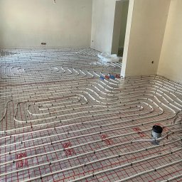 MJM-Instal Michał Matysik - Rozłożone rury ogrzewania podłogowego na folii z siatką, widoczne złączki i fragment ściany w surowym stanie.