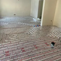 Rozłożone rury ogrzewania podłogowego na folii z siatką, widoczne złączki i fragment ściany w surowym stanie.