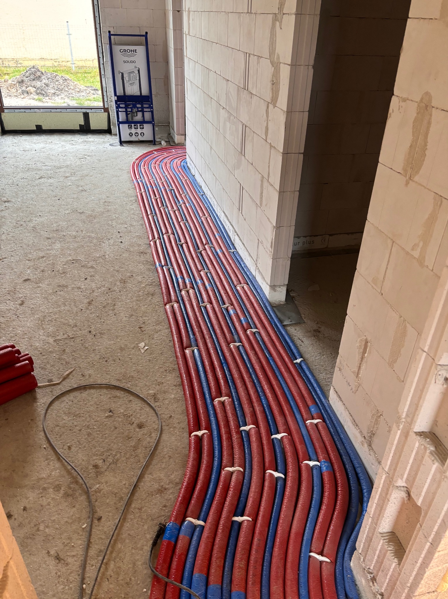 Równolegle ułożone, czerwono-niebieskie rury instalacyjne w surowym wnętrzu budynku z bloczków, widoczna rama podtynkowa Grohe i przewód elektryczny.