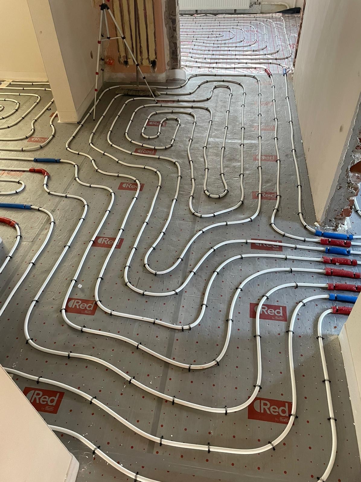 Rozłożona instalacja ogrzewania podłogowego z białych rur na folii z logo Red by Roth, widoczne złącza w kolorach czerwonym i niebieskim, surowy stan wykończenia pomieszczenia.