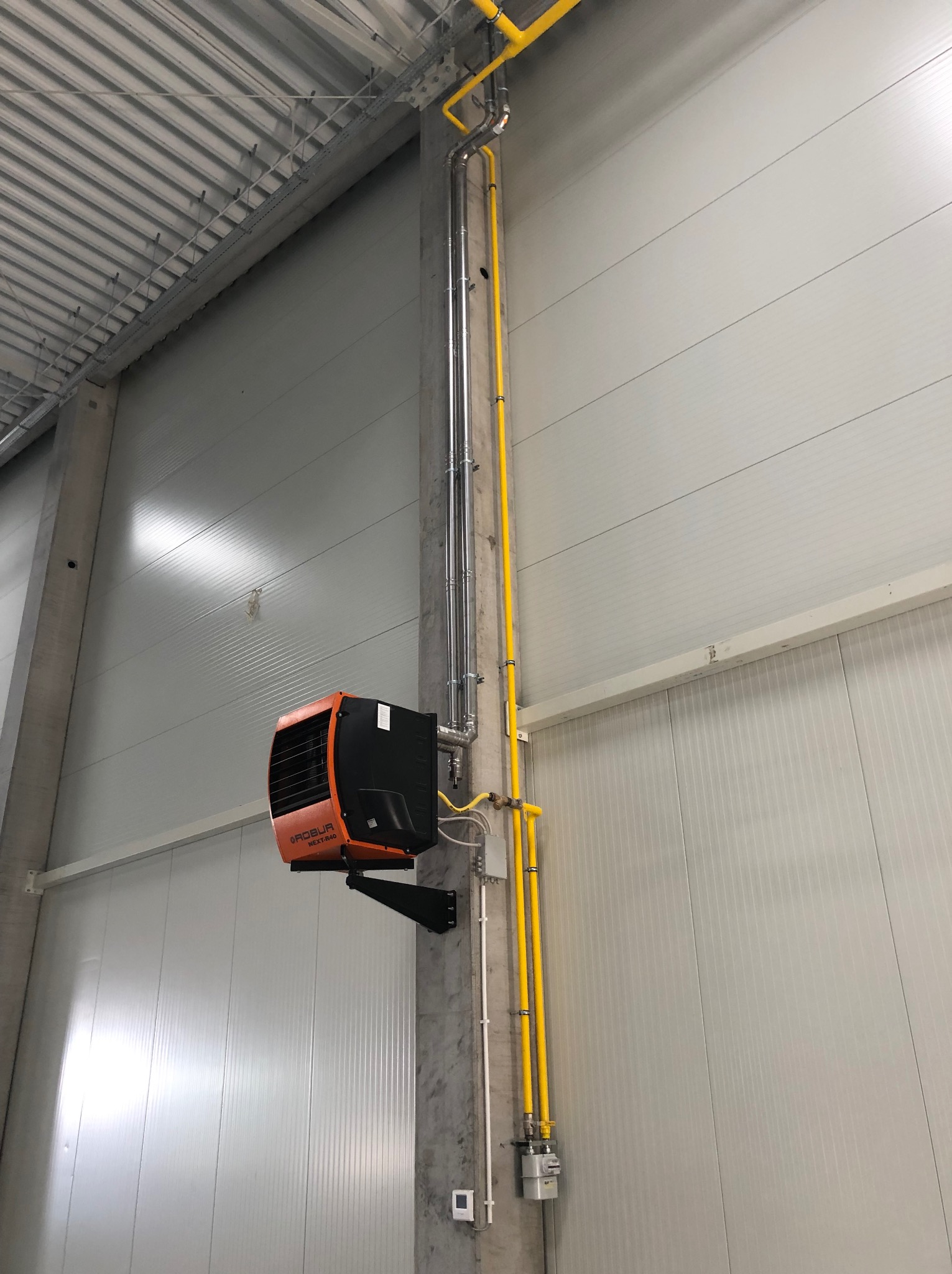 Zamontowany piec grzewczy Robur NEXT-R40 na betonowym słupie w hali, widoczne rury gazowe w kolorze żółtym i instalacja odprowadzająca spaliny, sufit z blachy trapezowej.