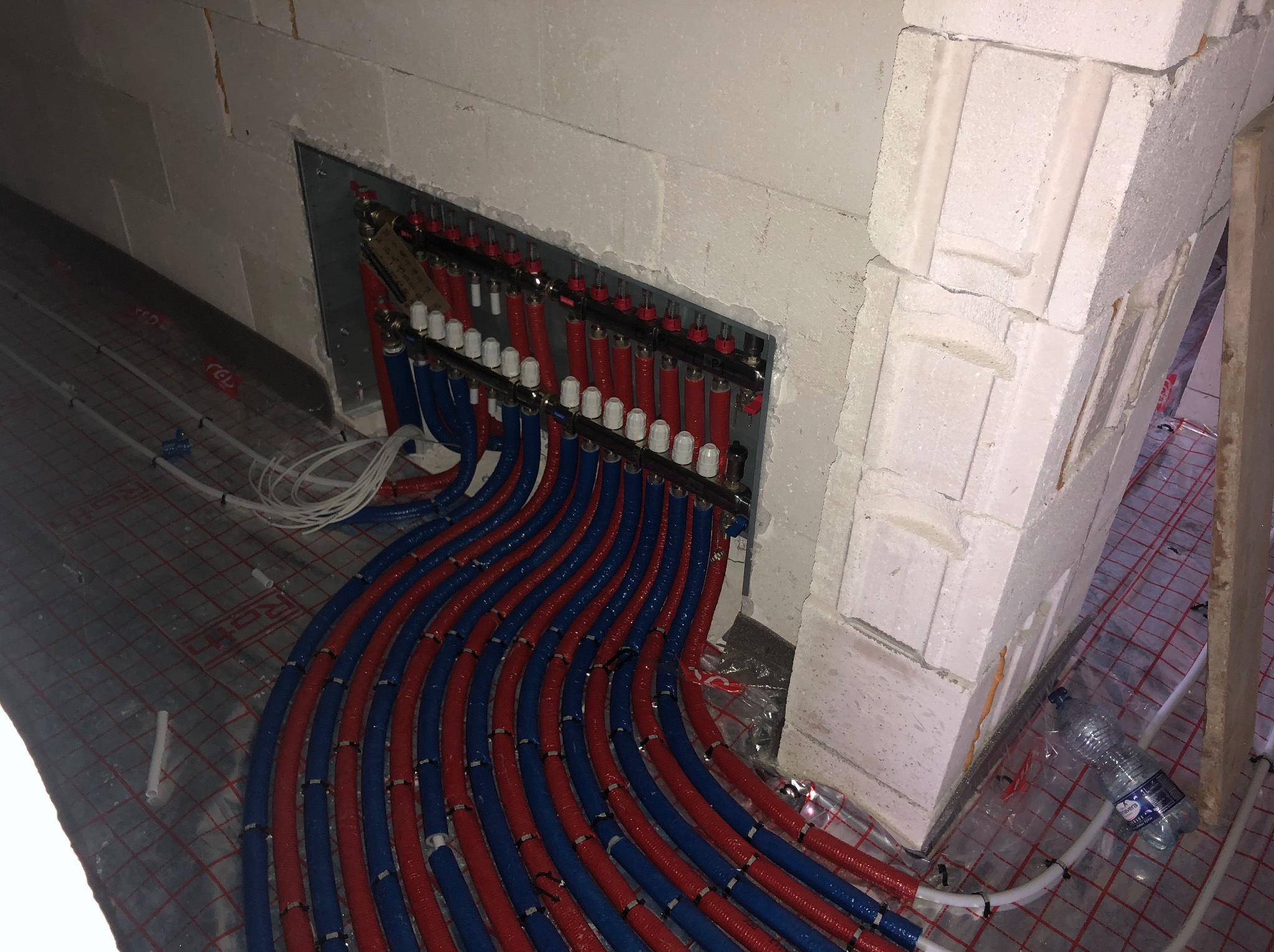 Rozdzielacz instalacji ogrzewania podłogowego z licznymi czerwonymi i niebieskimi rurami w nowym domu w Miasteczku Śląskim, widok z góry, widoczna butelka wody mineralnej w tle.