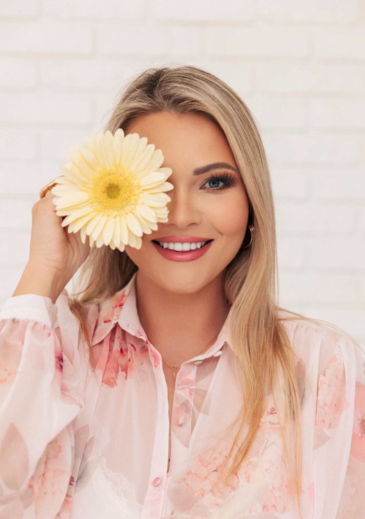 Uśmiechnięta blondynka z niebieskim okiem zasłoniętym żółtą gerberą, nosząca bluzkę w kwiaty i złote kolczyki koła na tle białej ceglanej ściany.