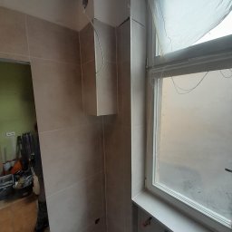 ArtDekor - Świeżo wykafelkowana łazienka z beżowymi płytkami, widoczne niedociągnięcia w postaci wystających kabli i niezamaskowanych otworów w ścianie, okno z zasłoniętą szybą i narzędzia w tle.