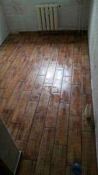 Ułożona podłoga z paneli imitujących drewno w pomieszczeniu z grzejnikiem i butelką płynu do czyszczenia w rogu. Widoczne niedoskonałości na powierzchni paneli.