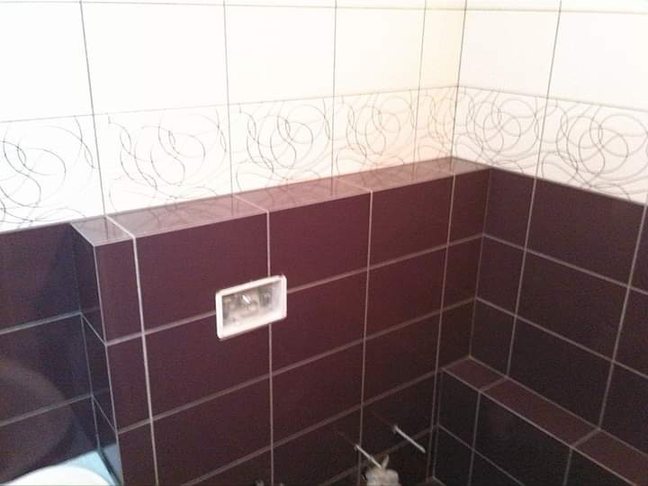 Wykończona ściana w łazience z bordowymi płytkami i białym pasem z wzorem, widoczny otwór na przycisk spłuczki.