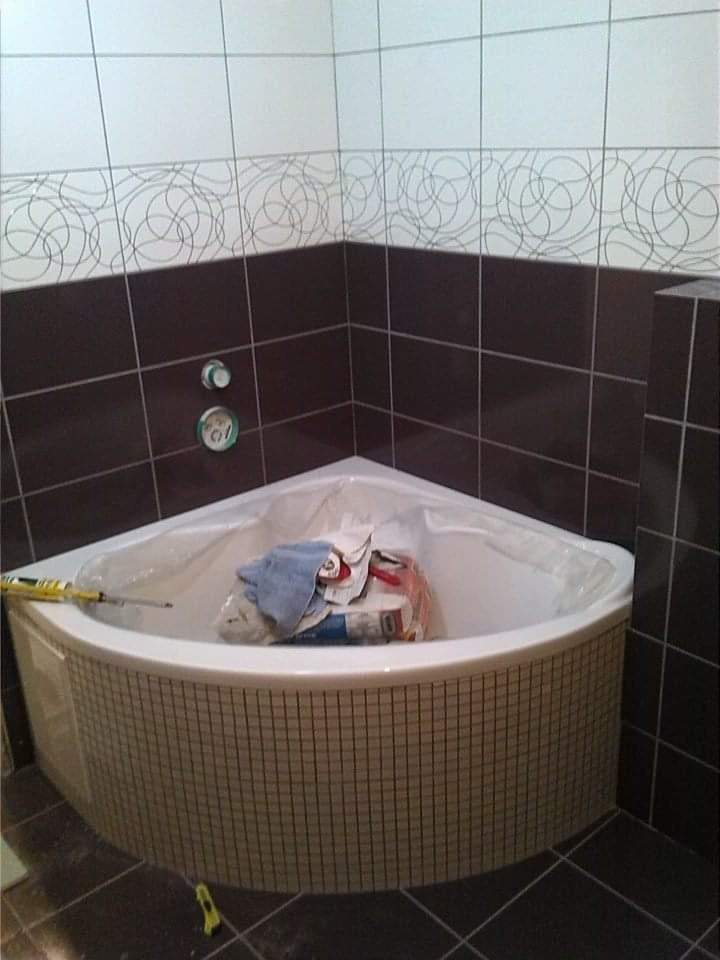 Narożna wanna z hydromasażem zabezpieczona folią, otoczona brązowymi i białymi płytkami z motywem okręgów, wewnątrz wanny leżą narzędzia i materiały budowlane.