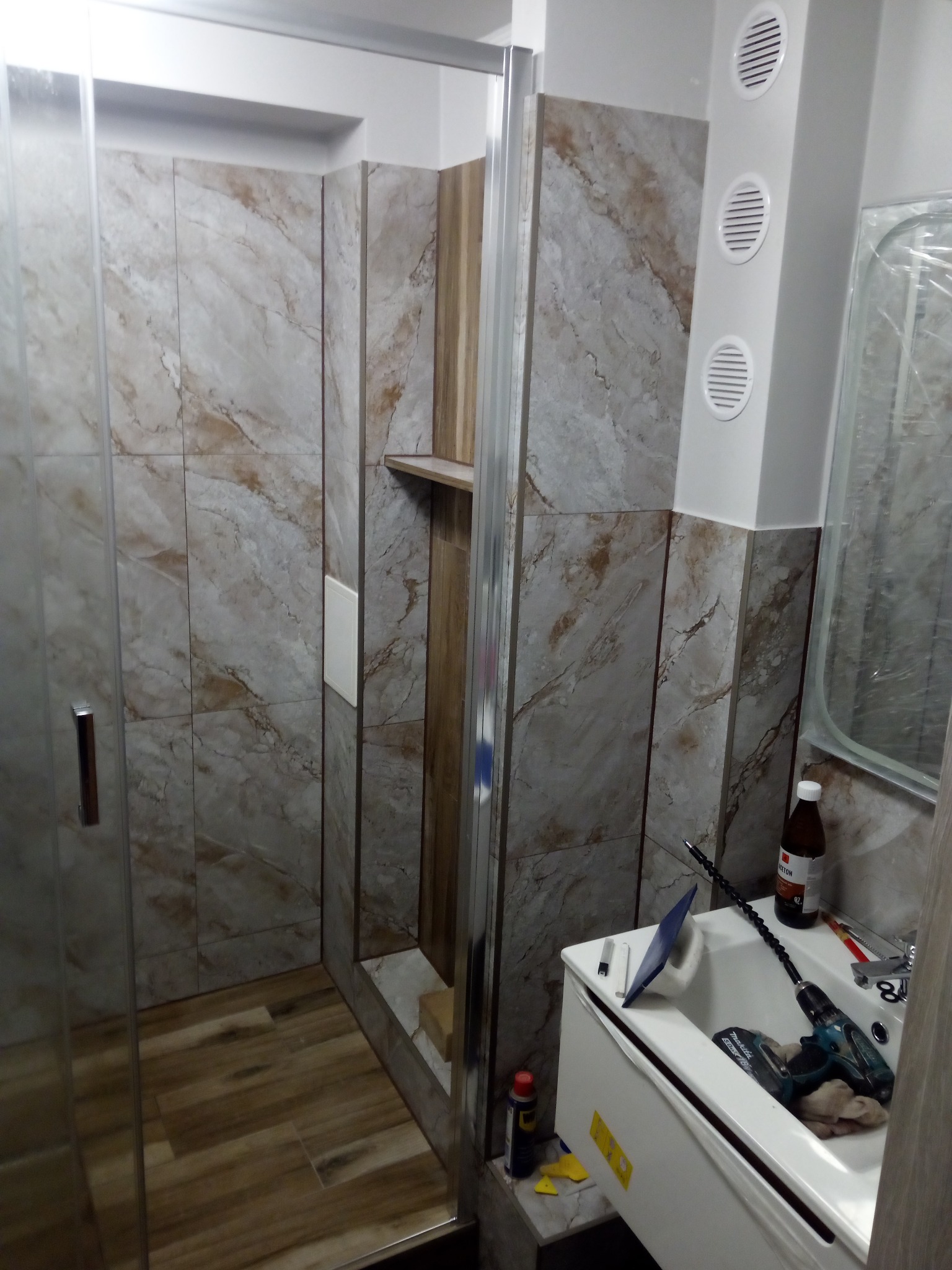 Ukończona kabina prysznicowa z marmurkowymi płytkami w odcieniach beżu i szarości, widoczna umywalka z narzędziami i akcesoriami do montażu, w tle trzy okrągłe kratki wentylacyjne na pionowym panelu.