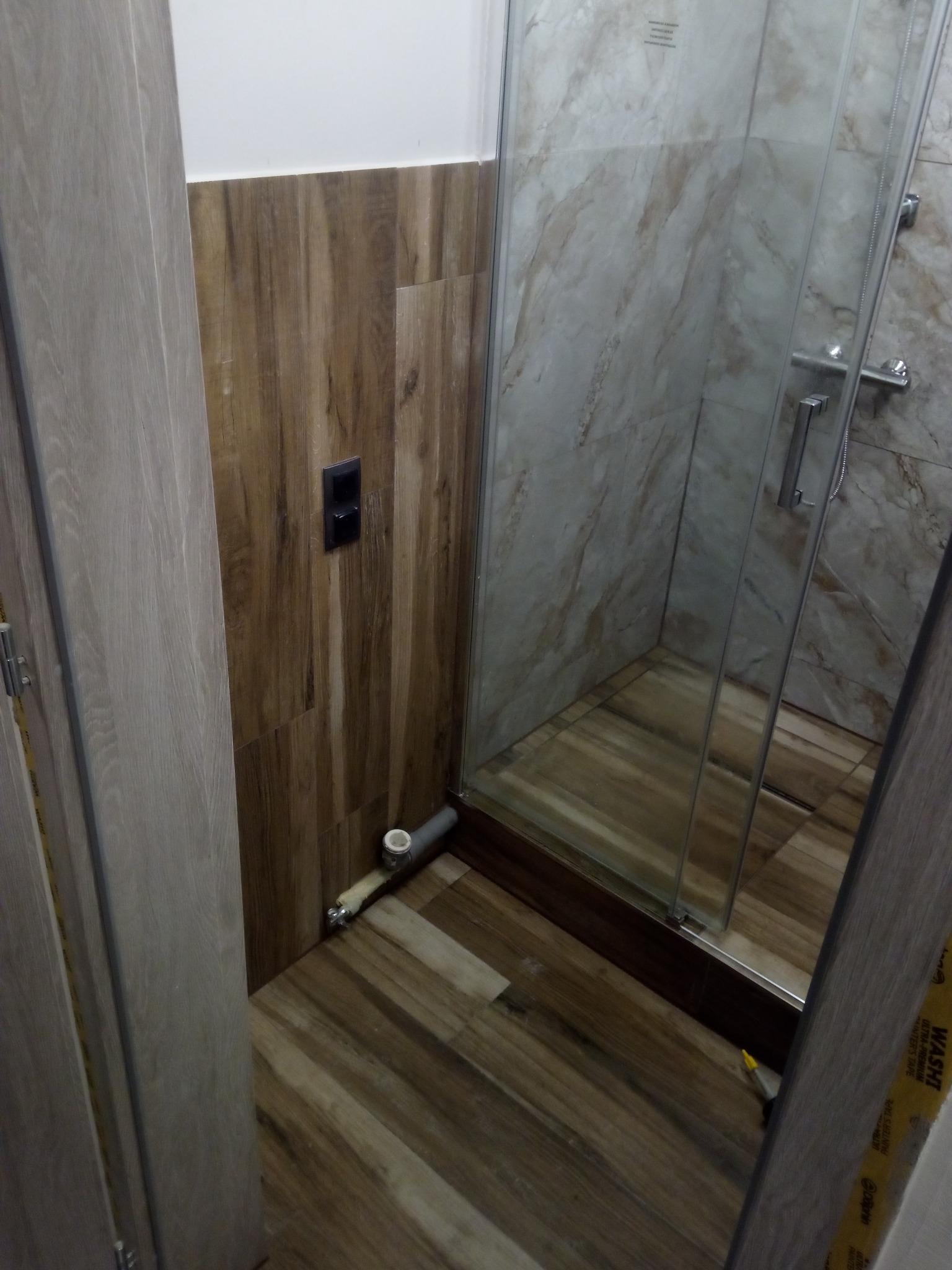 Fragment łazienki w trakcie remontu, widoczna kabina prysznicowa z marmurowymi płytkami, drewnopodobne panele na ścianie i podłodze, oraz wystające rury odpływowe przed montażem. Na podłodze leży...