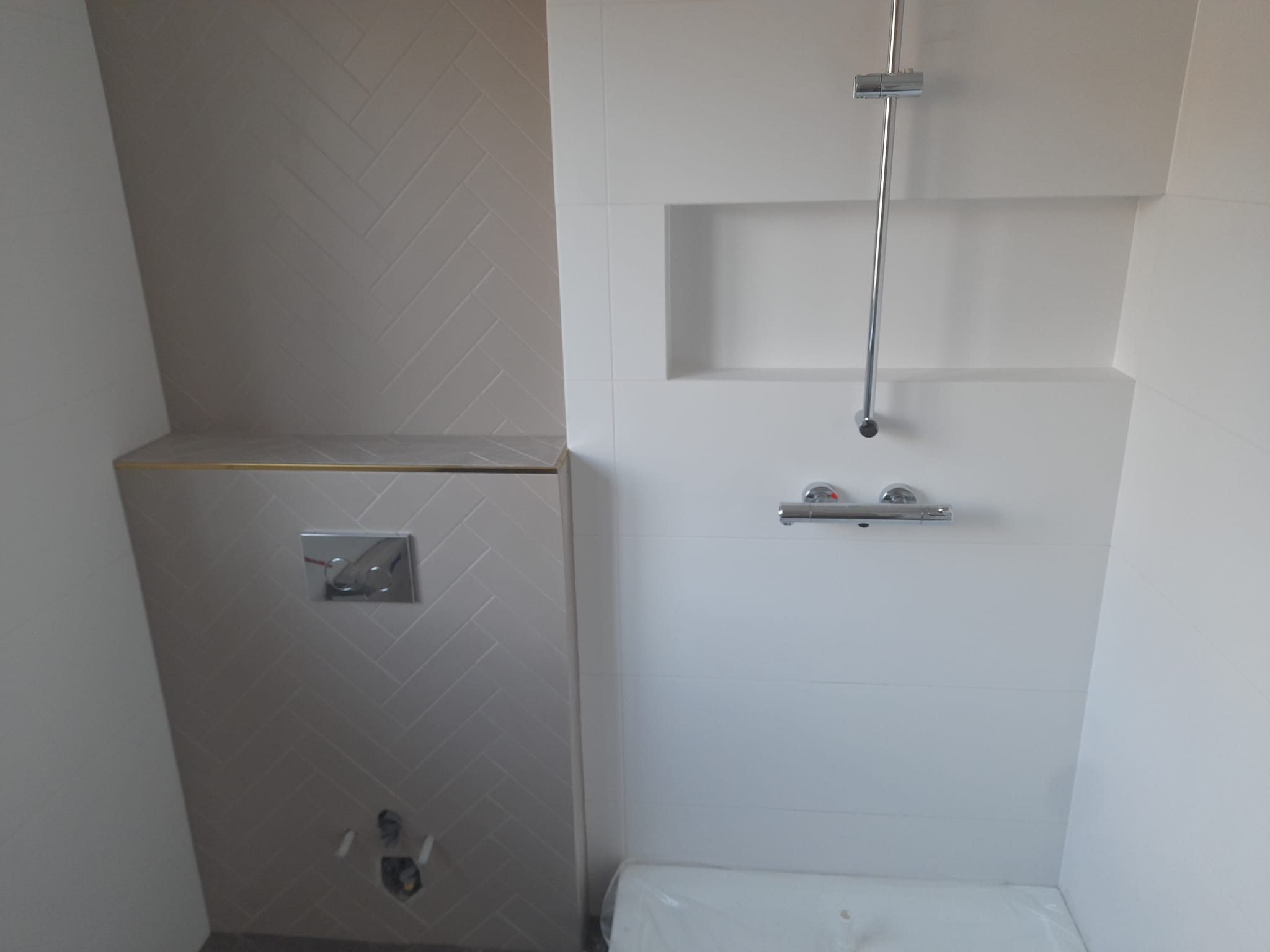 Nowoczesna łazienka z kremowymi płytkami ułożonymi w jodełkę na ścianie obok stelaża WC, minimalistyczna armatura prysznicowa i wnęka w ścianie wykończona białymi płytkami.