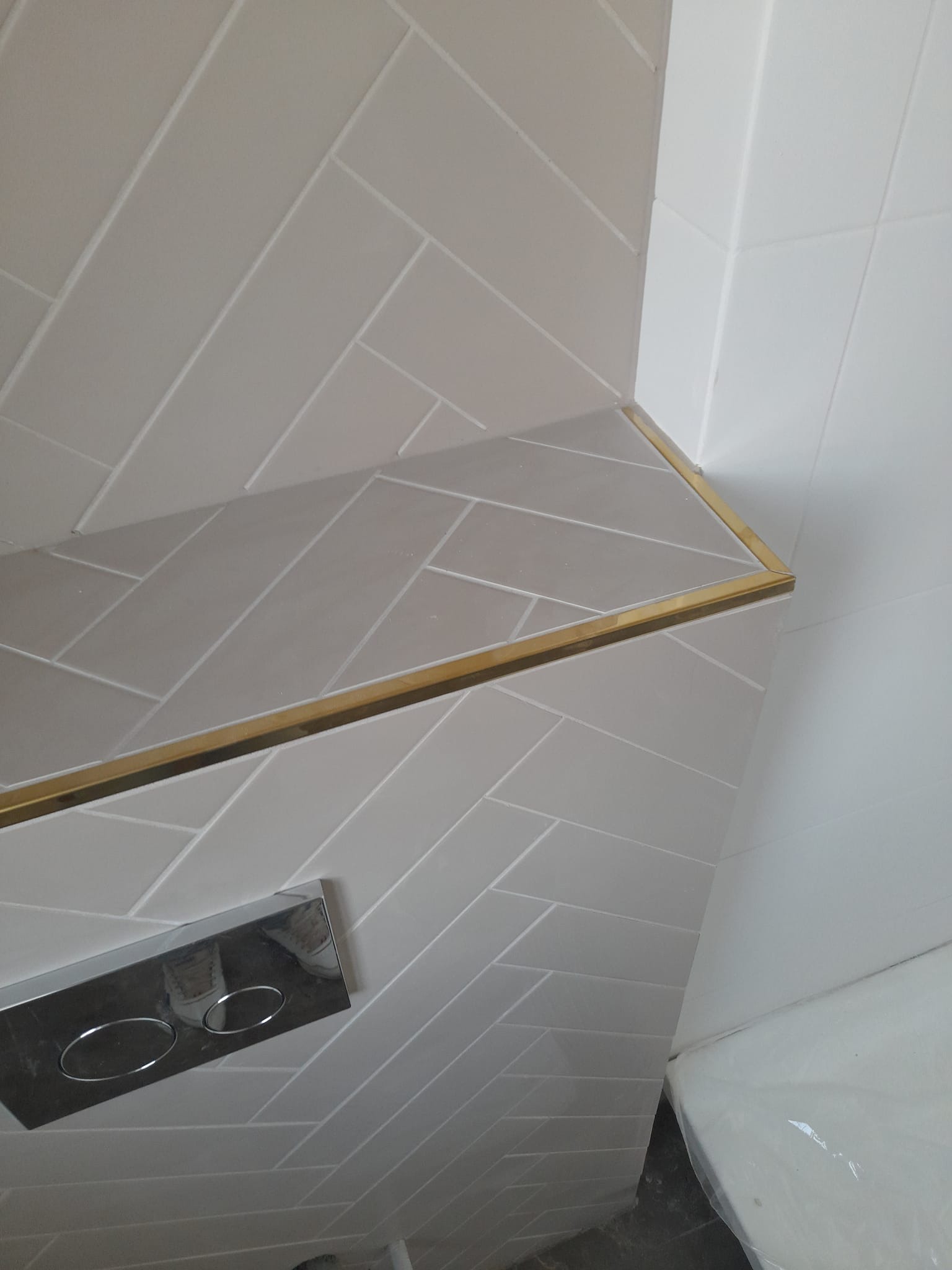 Wykończona półka w łazience z jasnych, ułożonych w jodełkę płytek ceramicznych, wykończona złotą listwą, widoczny przycisk spłuczki i fragment toalety.