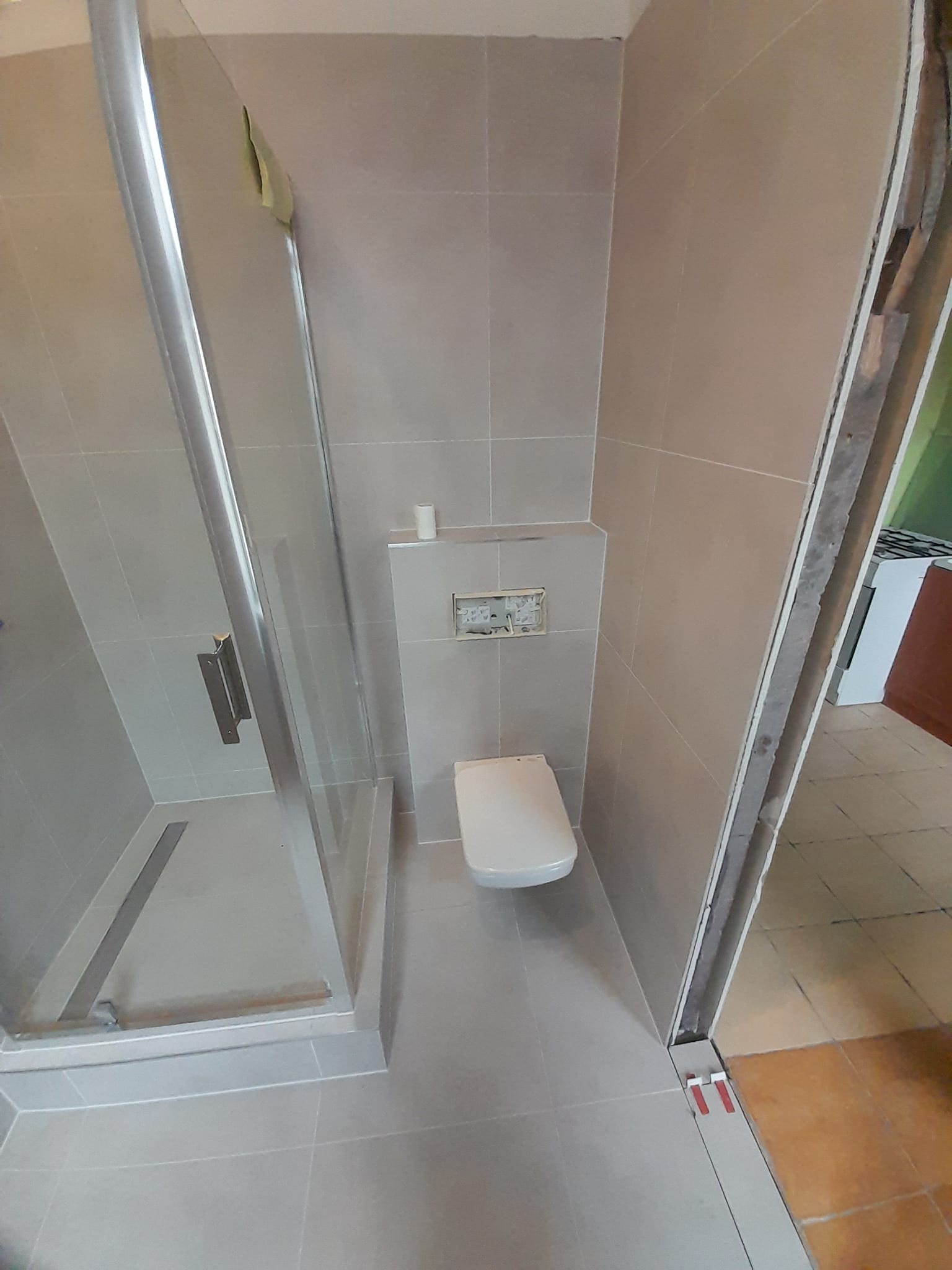 Zamontowana kabina prysznicowa z odpływem liniowym i podwieszana toaleta w łazience wykończonej jasnymi, kwadratowymi płytkami. Widoczne elementy instalacji podtynkowej. Fragment otwartych drzwi...