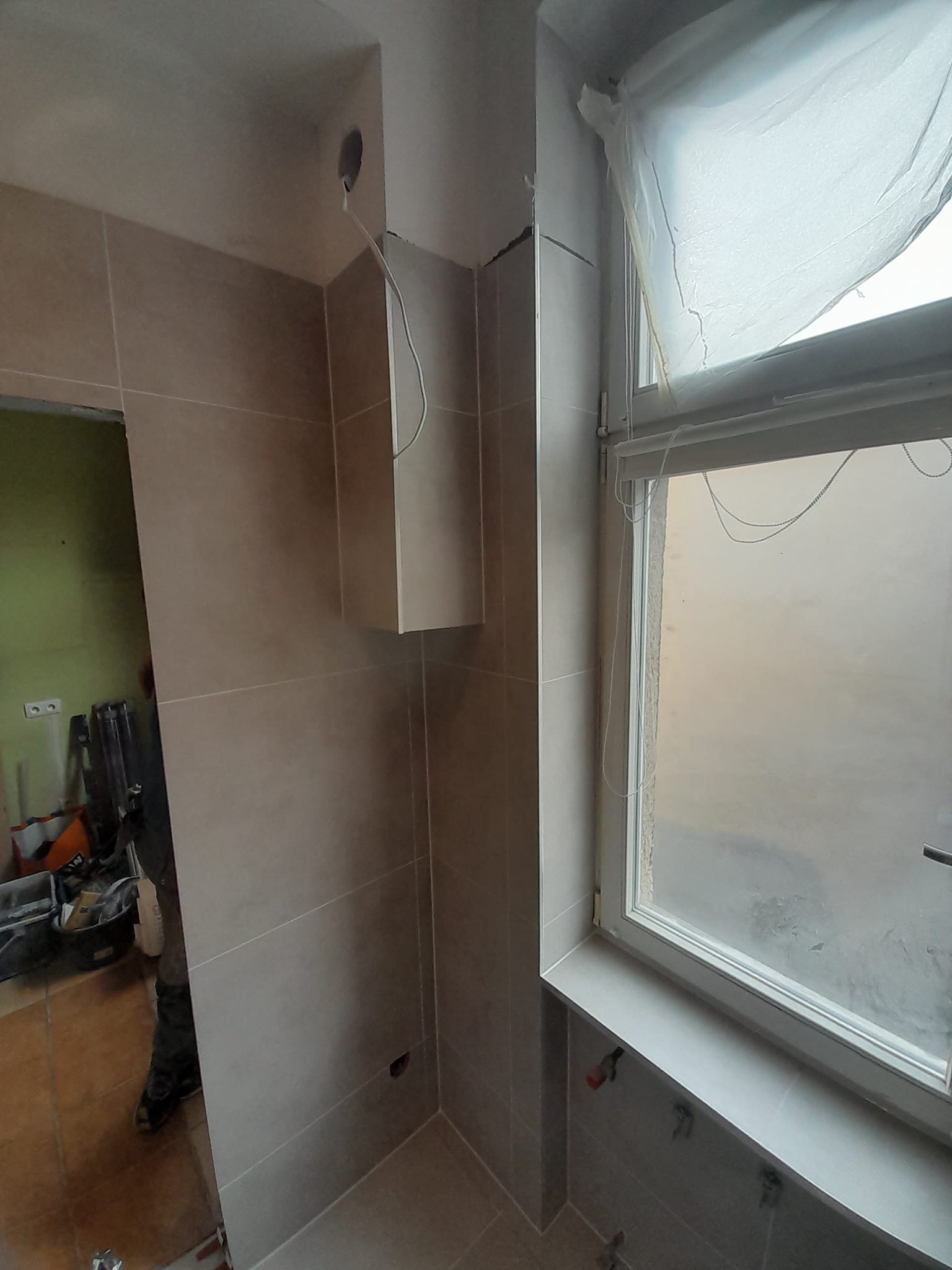 Świeżo wykafelkowana łazienka z beżowymi płytkami, widoczne niedociągnięcia w postaci wystających kabli i niezamaskowanych otworów w ścianie, okno z zasłoniętą szybą i narzędzia w tle.