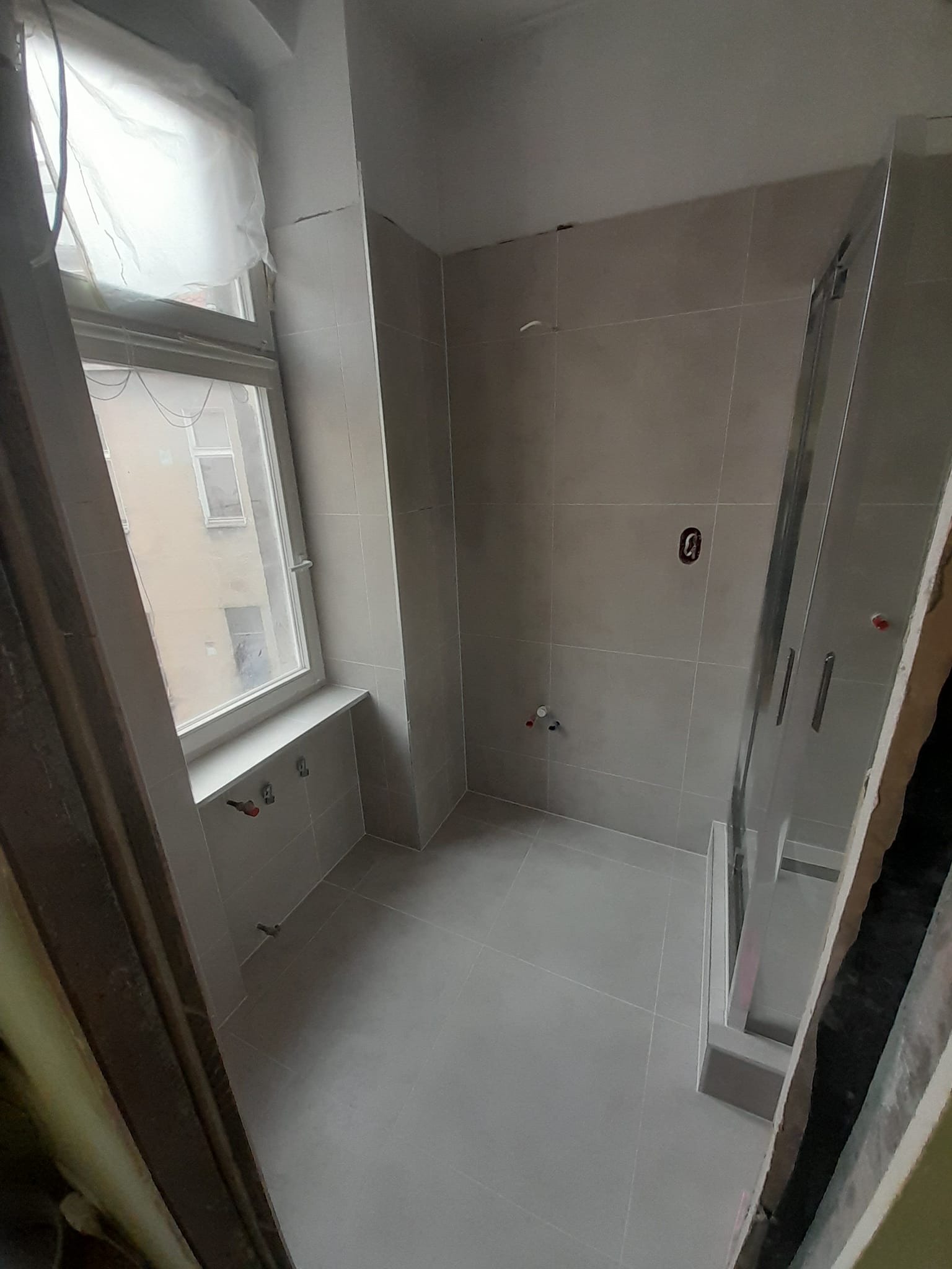 Wnętrze łazienki w trakcie remontu, widoczne szare płytki na ścianach i podłodze, zamontowana kabina prysznicowa z chromowanymi uchwytami, zabezpieczone okno folią malarską, wystające rury...