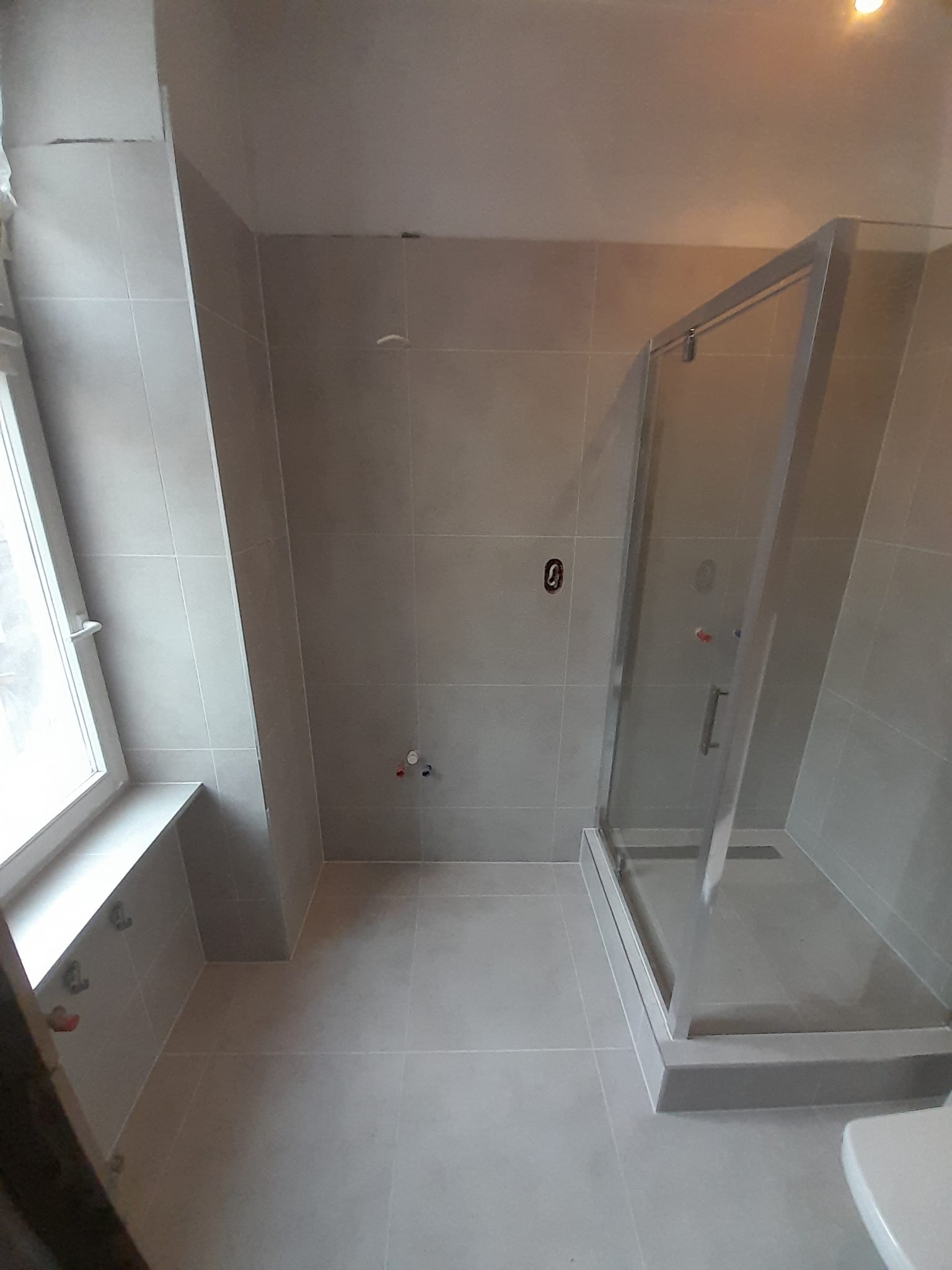 Nowoczesna łazienka z szarymi płytkami, kabiną prysznicową ze srebrnymi ramami i oknem z białymi ramami, widoczne wystające ze ściany przyłącza wodne.