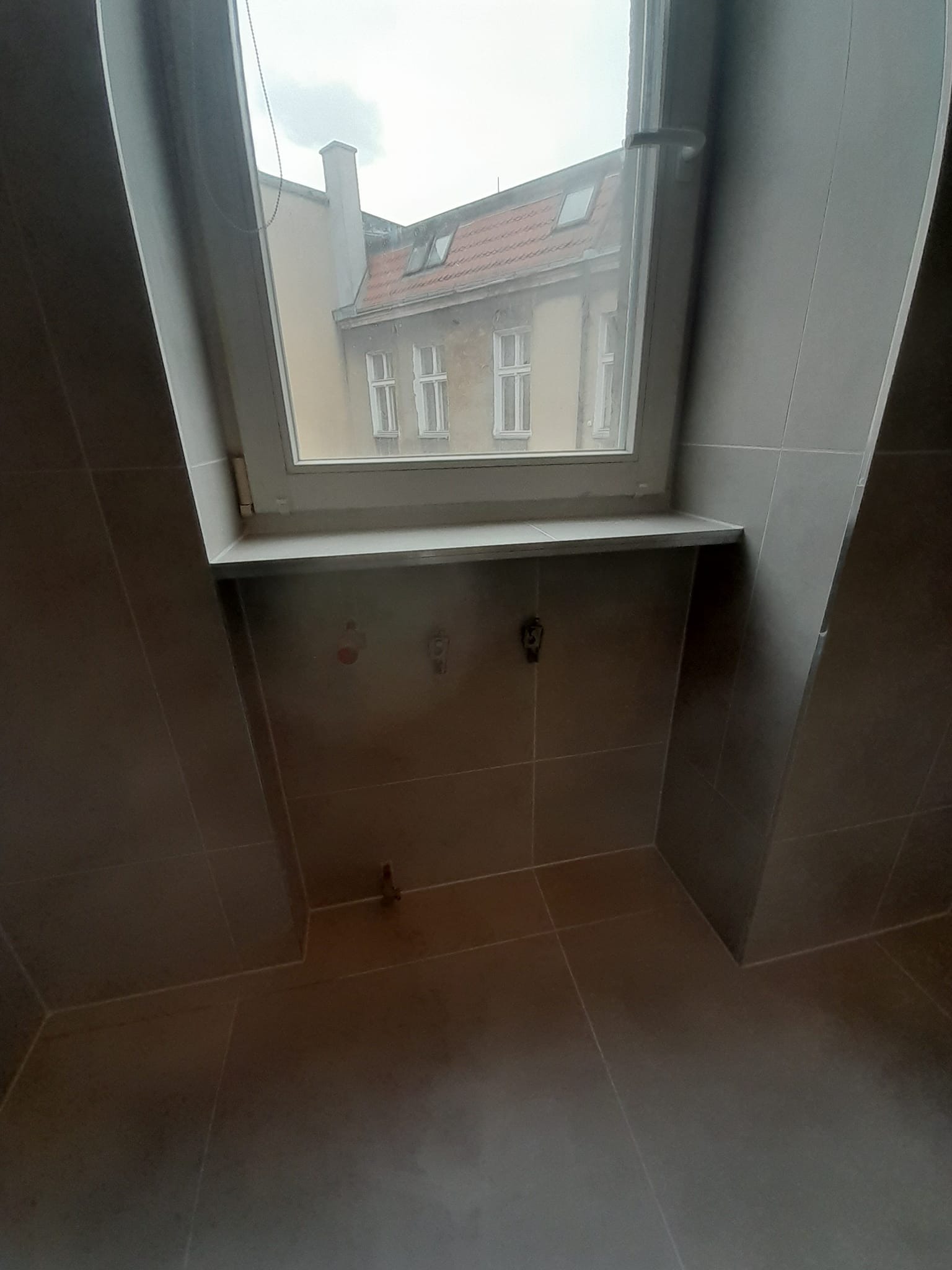 Szara łazienka z oknem dachowym, widoczne instalacje wodne na ścianie, płytki ceramiczne na ścianach i podłodze, budynek z czerwonym dachem za oknem.