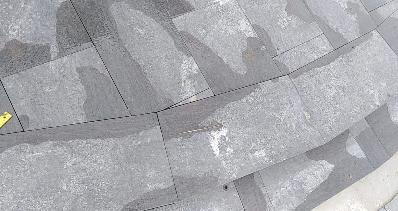 Szare, prostokątne płytki ułożone na schodach, widoczne ślady wilgoci i żółta miarka w lewym dolnym rogu.