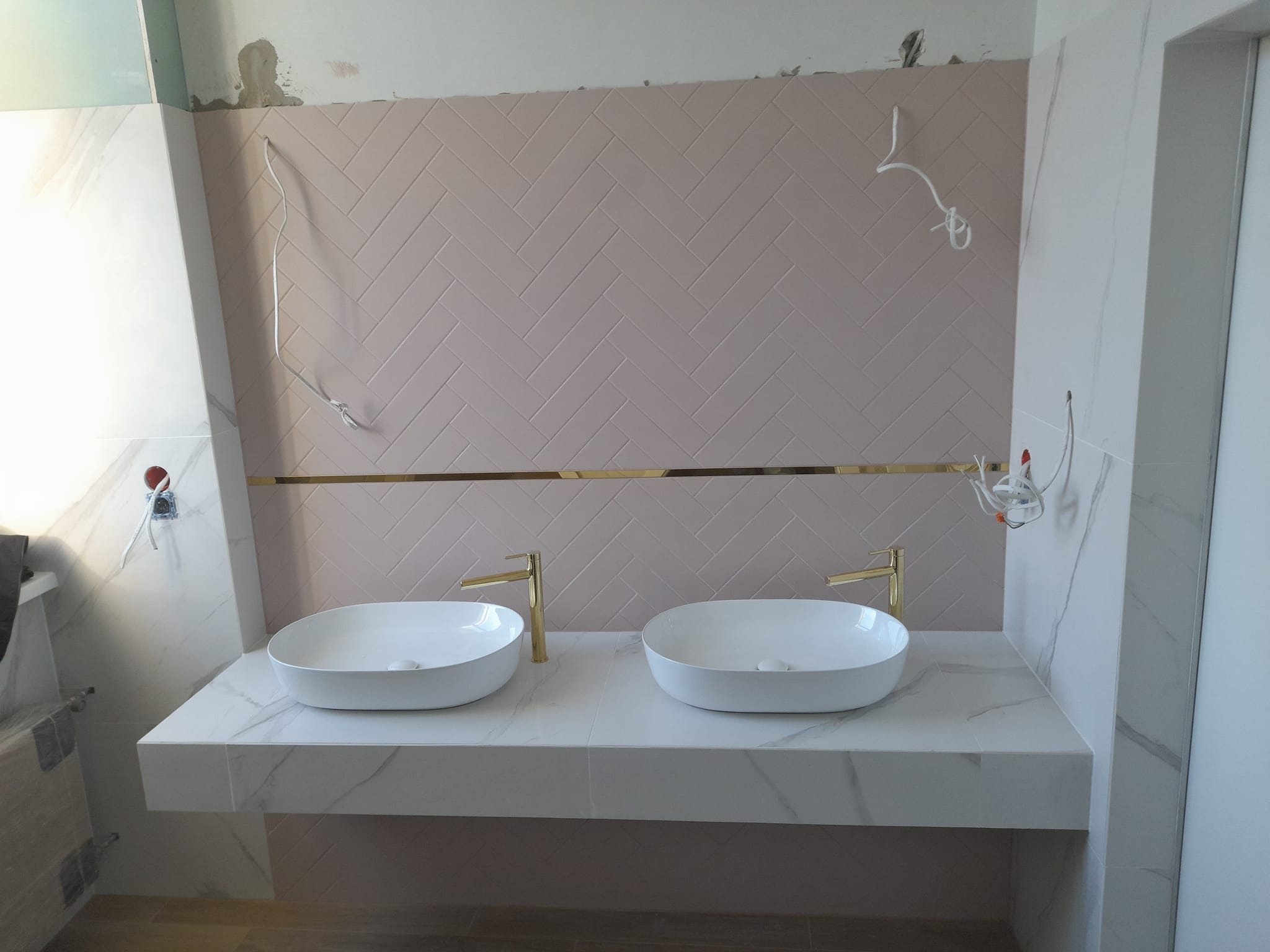 Niedokończona łazienka z dwoma białymi umywalkami nablatowymi, złotą armaturą i ścianą wyłożoną różową glazurą w jodełkę, widoczne przewody elektryczne i niezakończone prace wykończeniowe.