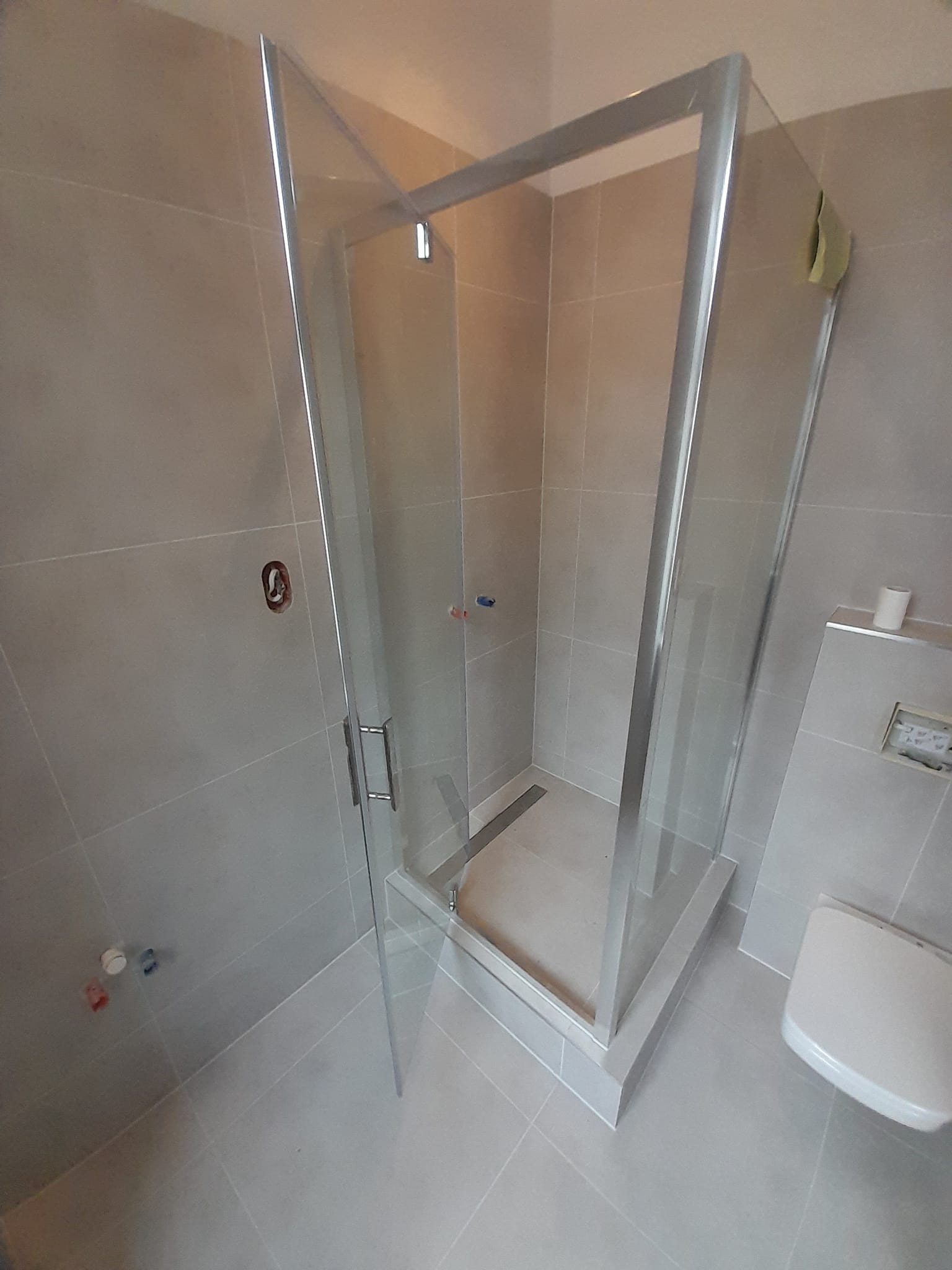 Szklana kabina prysznicowa z metalową ramą w łazience wyłożonej szarymi płytkami, widoczne przyłącza wodne i toaleta podtynkowa