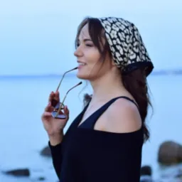 Uśmiechnięta brunetka w chustce w panterkę, trzymająca okulary przeciwsłoneczne na tle błękitnego morza.